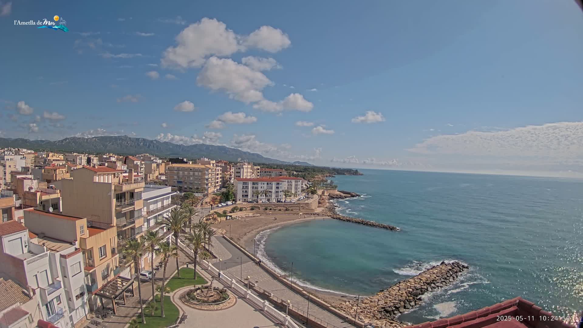 A coastal town with buildings and palm trees stretches along a calm, blue ocean under a mostly sunny sky with scattered clouds and distant mountains.