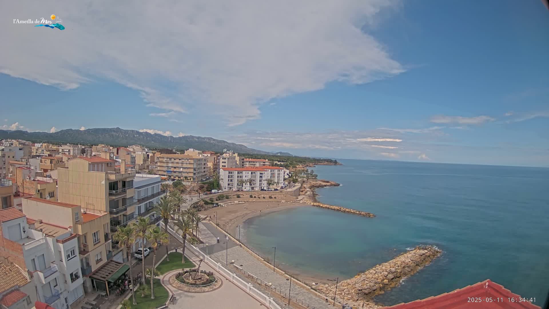 An aerial view shows a coastal town on a partly cloudy day, with a calm blue sea and a small sandy cove.