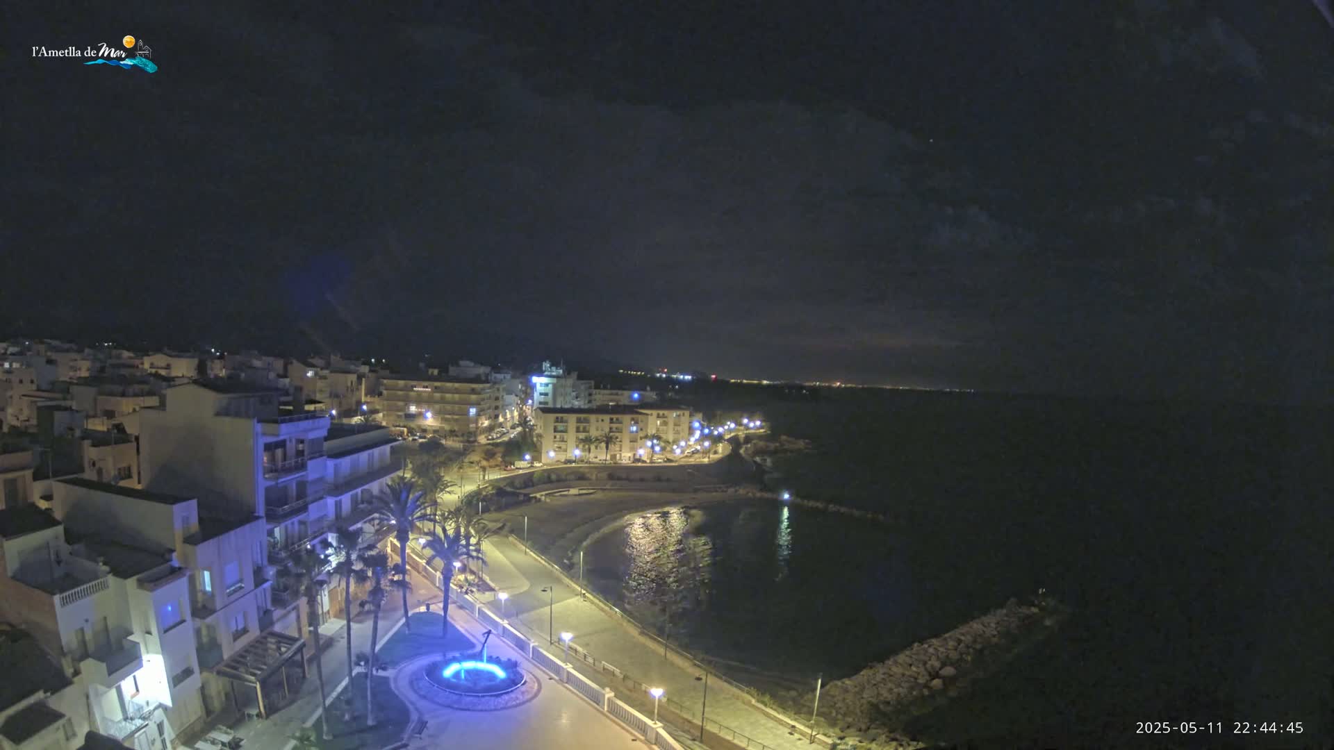 A nighttime coastal town is illuminated, showing a calm sea under a mostly clear sky.