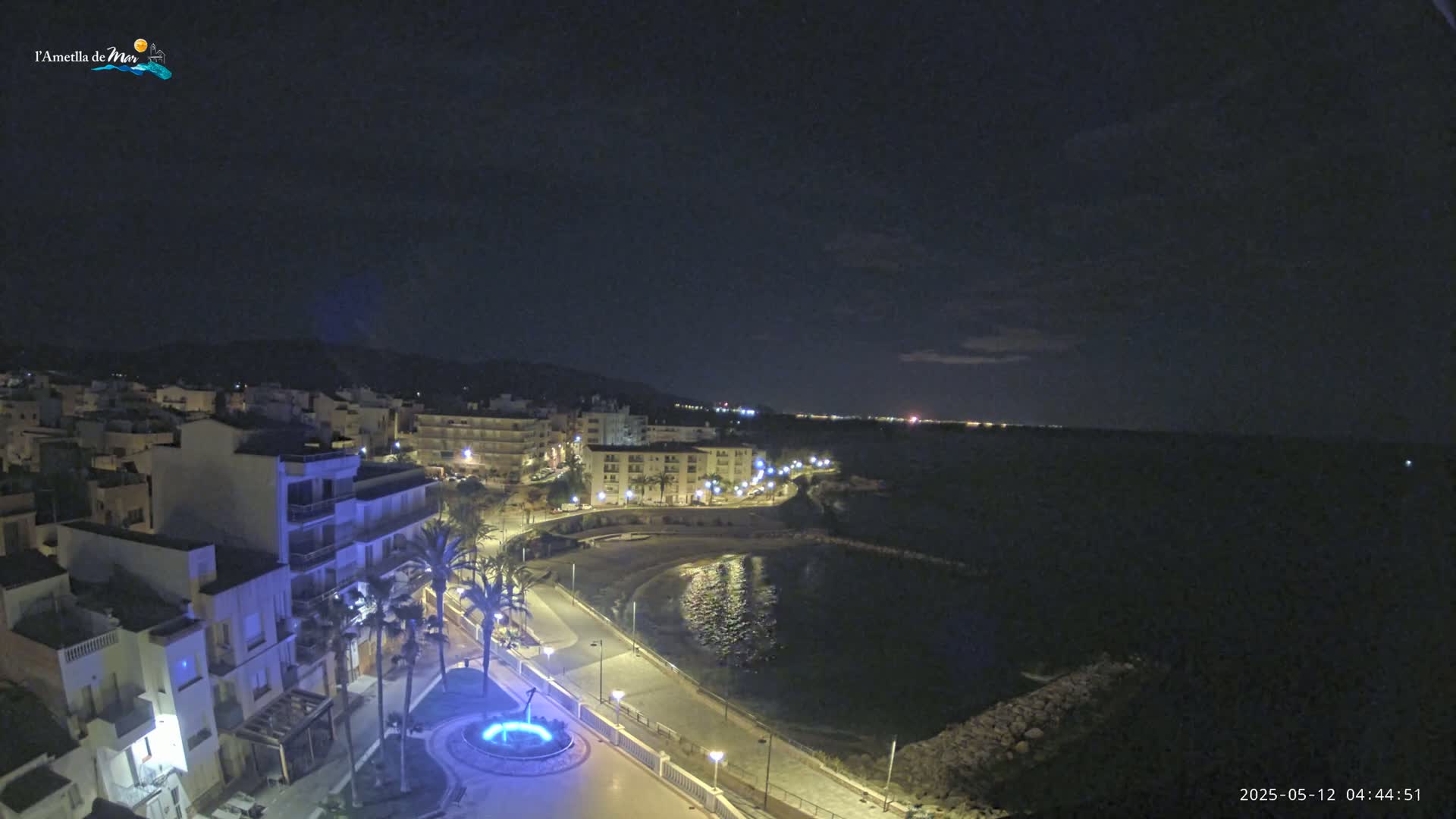 A nighttime coastal town scene shows a crescent-shaped beach and calm water under a mostly clear sky, with buildings and palm trees illuminated along a paved waterfront path.