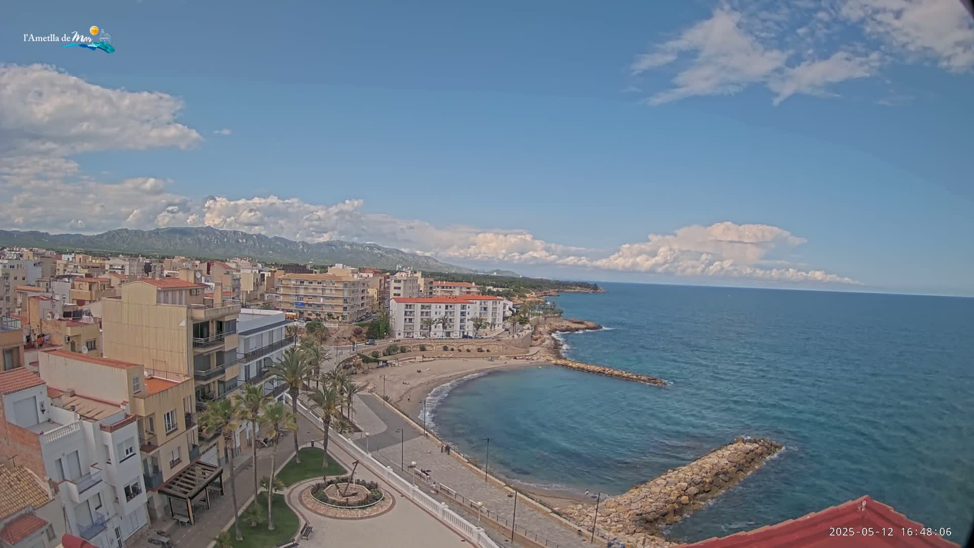 A coastal town with buildings, palm trees, a small sandy beach, and a calm blue sea under a partly cloudy sky.
