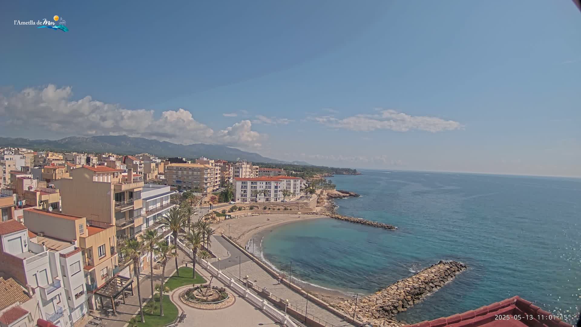 A coastal town with a small, sandy beach curving into a calm, blue ocean under a mostly sunny sky with scattered clouds and mountains in the distance.