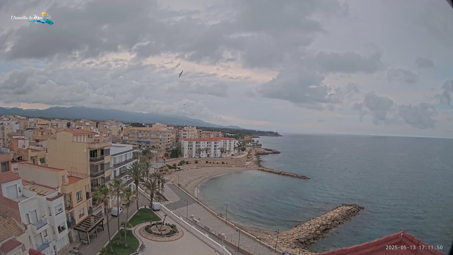 A coastal town with buildings, a small beach, and calm, cloudy waters extends to mountains in the distance under an overcast sky.