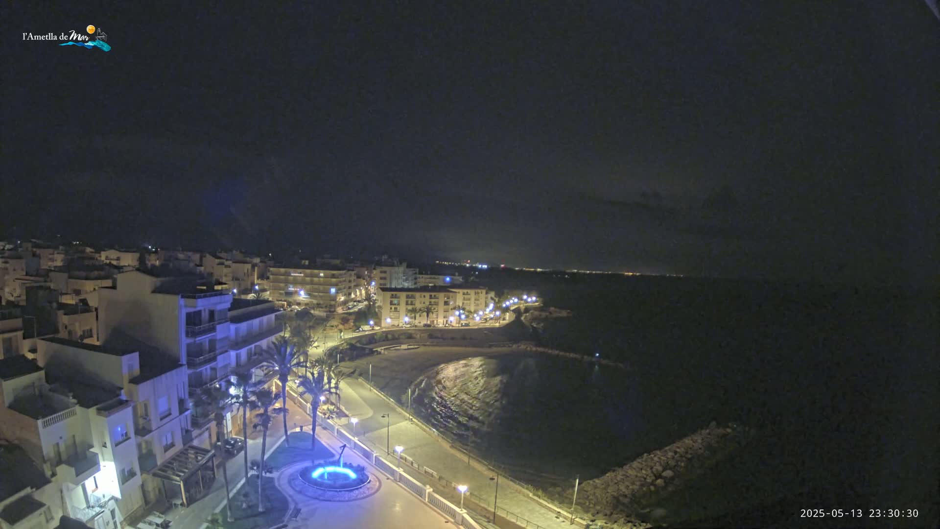 A nighttime view of a coastal town, with a crescent-shaped bay and a promenade illuminated by streetlights.