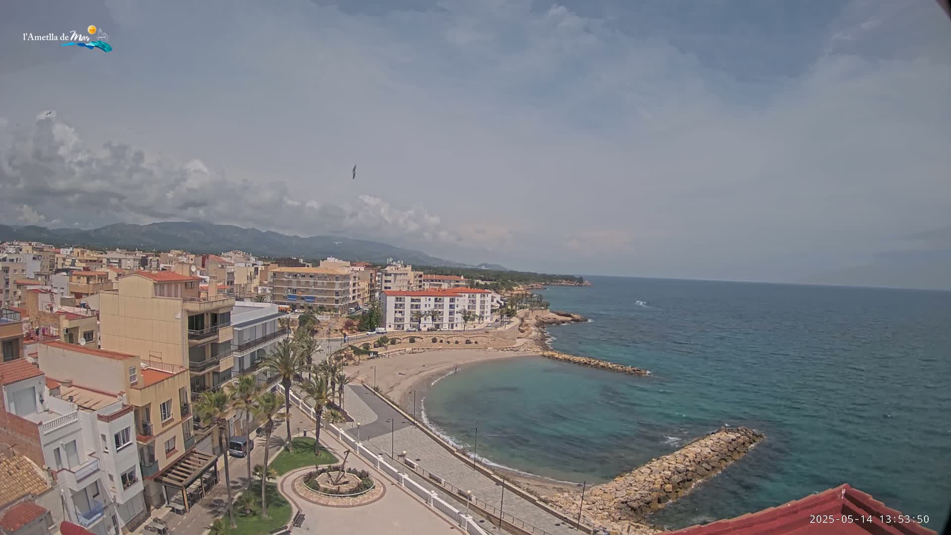 A partly cloudy day shows a coastal town with a small, curved beach and calm, teal water, backed by mountains in the distance.