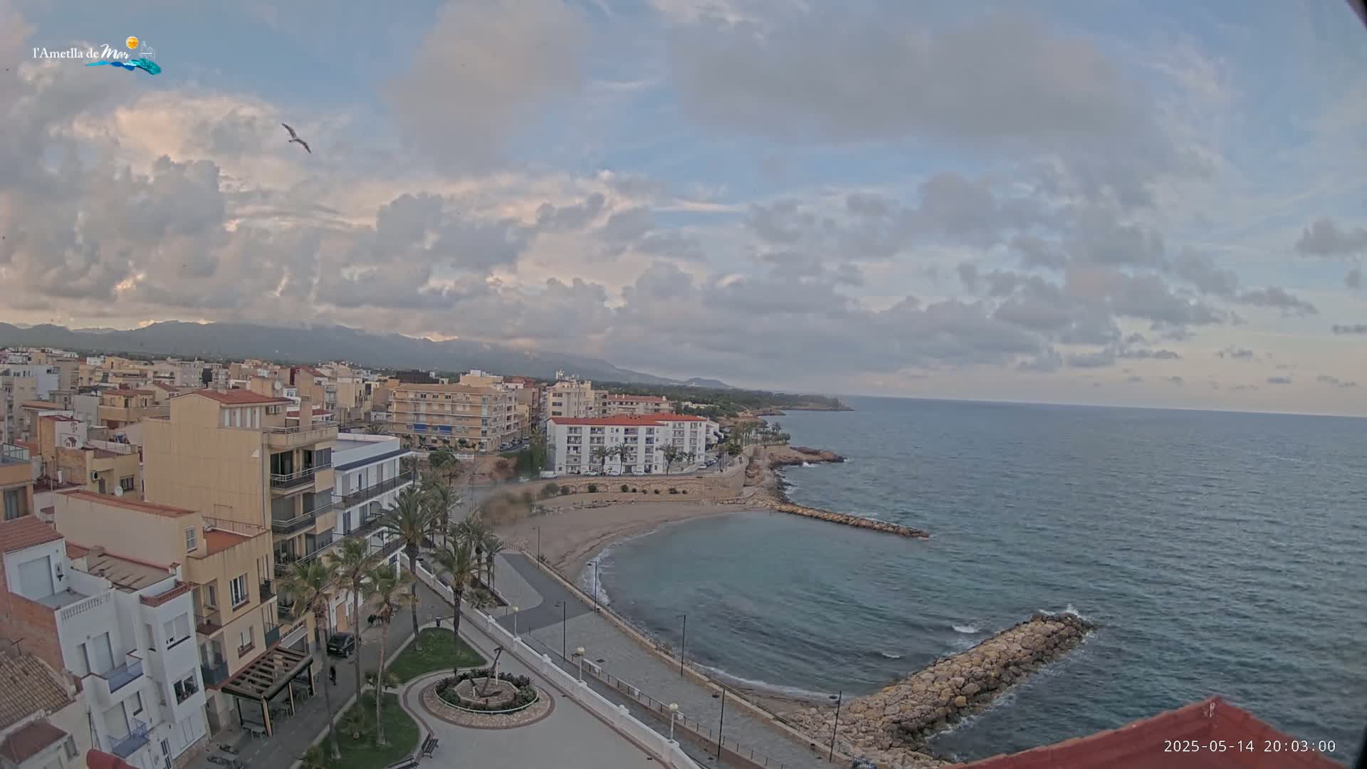 An aerial view shows a coastal town with buildings and a small sandy beach under a partly cloudy sky.