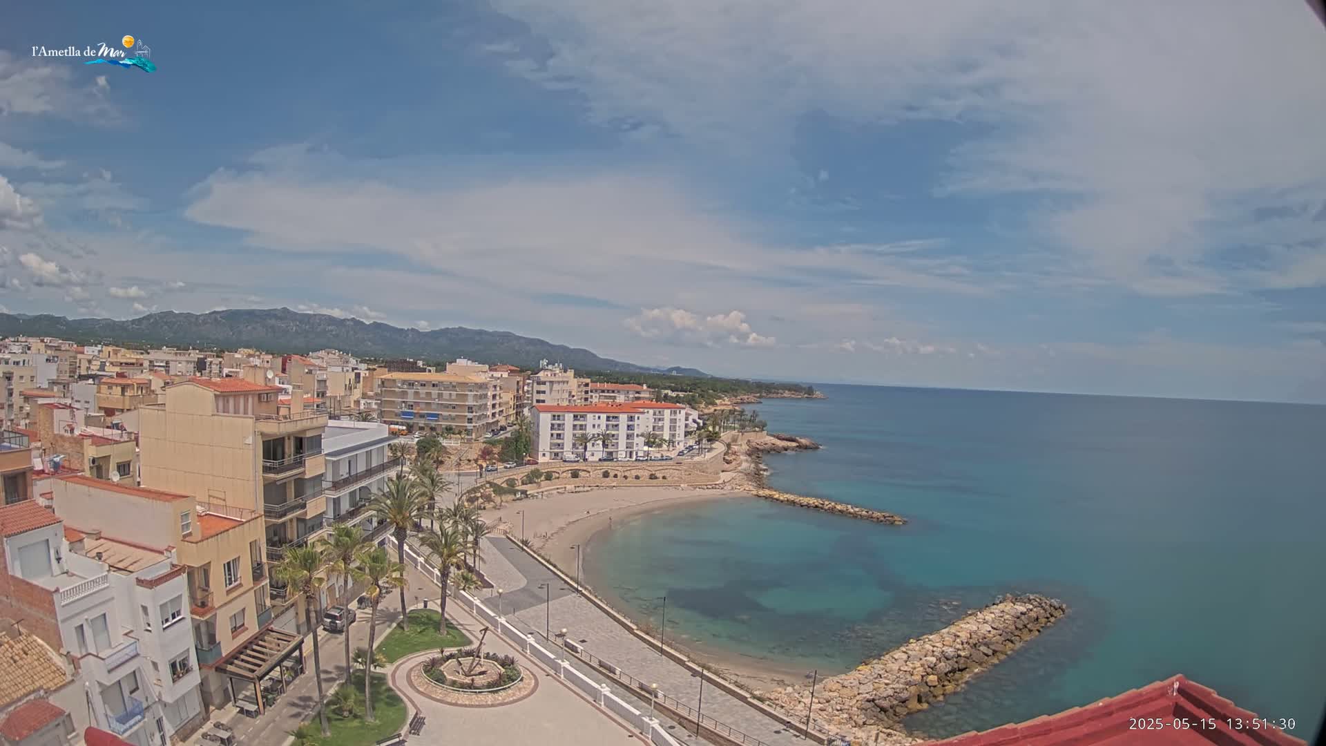 An elevated view shows a coastal town on a partly cloudy day, with a small sandy beach curving into a calm, teal sea partially enclosed by a rock jetty.