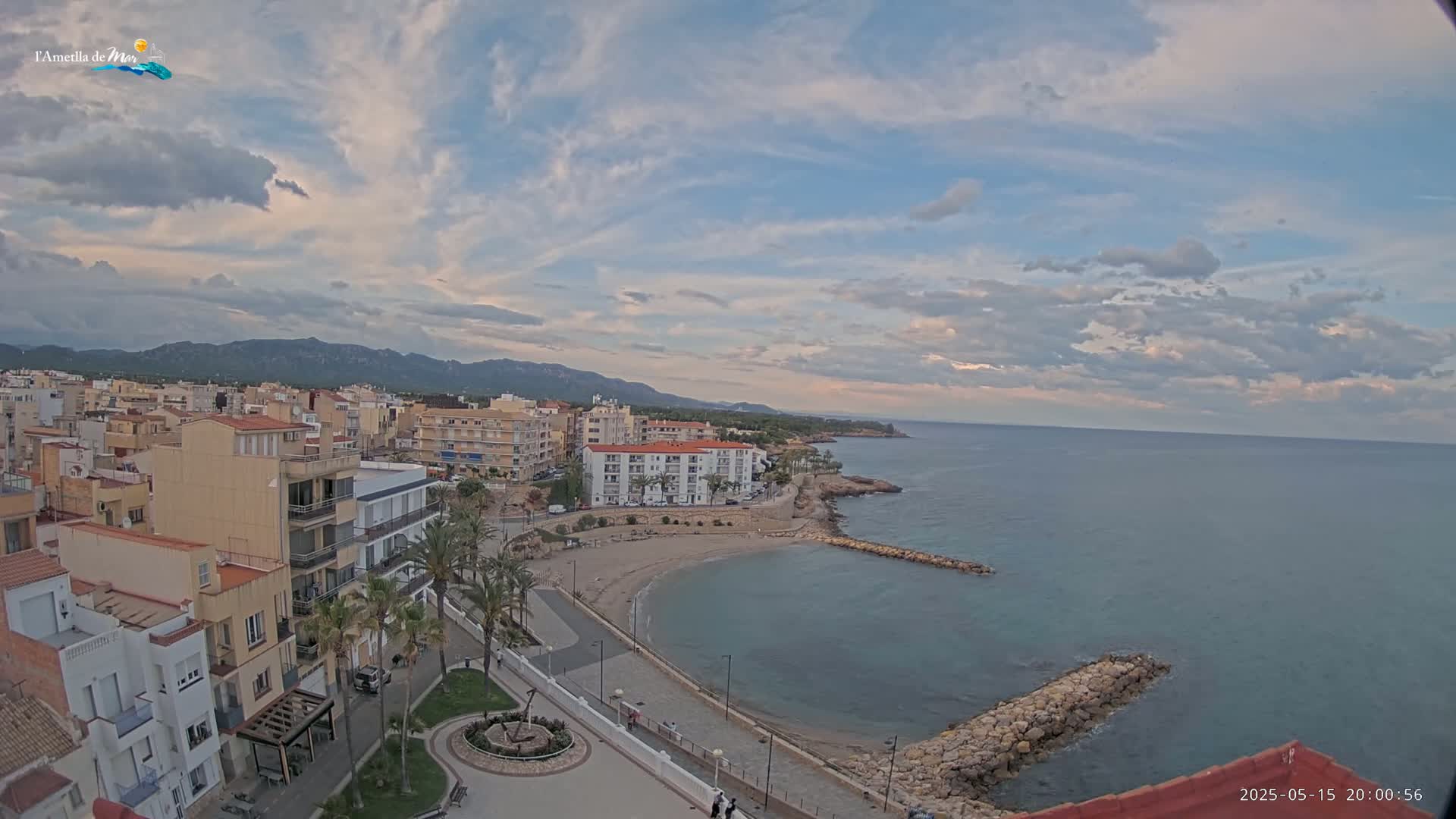 An elevated view of a coastal town under a partly cloudy sky shows a small, crescent-shaped beach curving into a calm sea, bordered by buildings and a rocky breakwater.