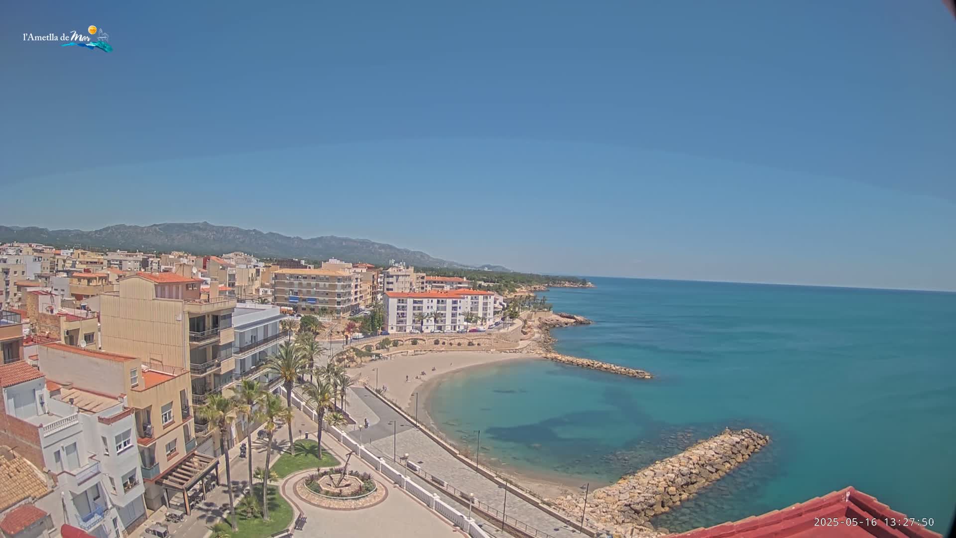 A coastal town with buildings, palm trees, and a small sandy beach curves into a calm, teal ocean under a clear blue sky.