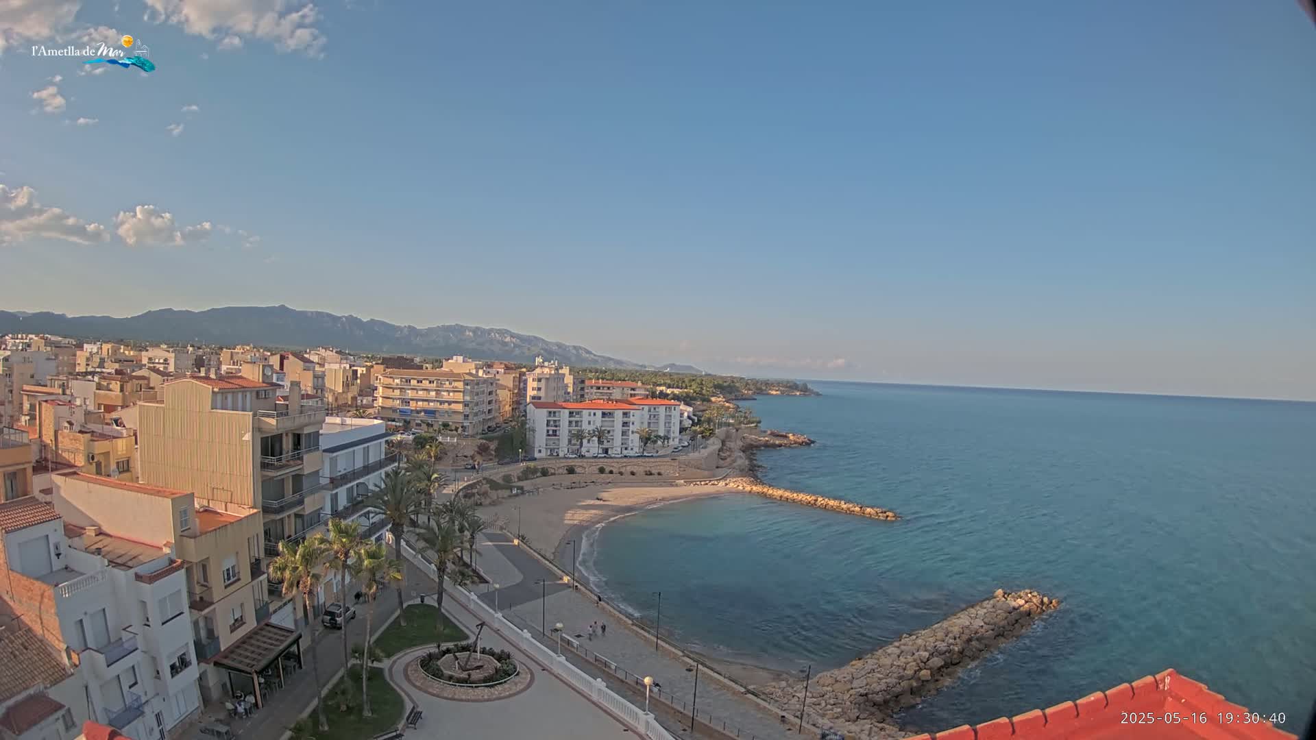 A coastal town with a small beach and calm, blue sea is seen under a mostly clear sky, with mountains in the background.