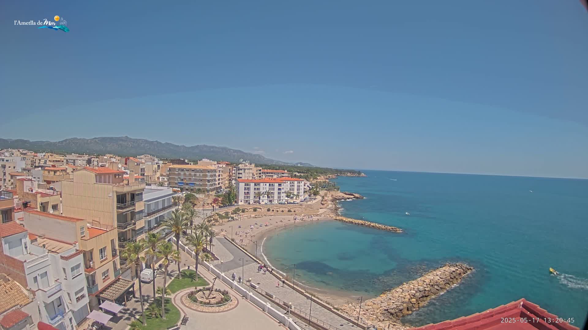 A coastal town with buildings, palm trees, and a small sandy beach curving into a clear turquoise sea under a bright, sunny sky.