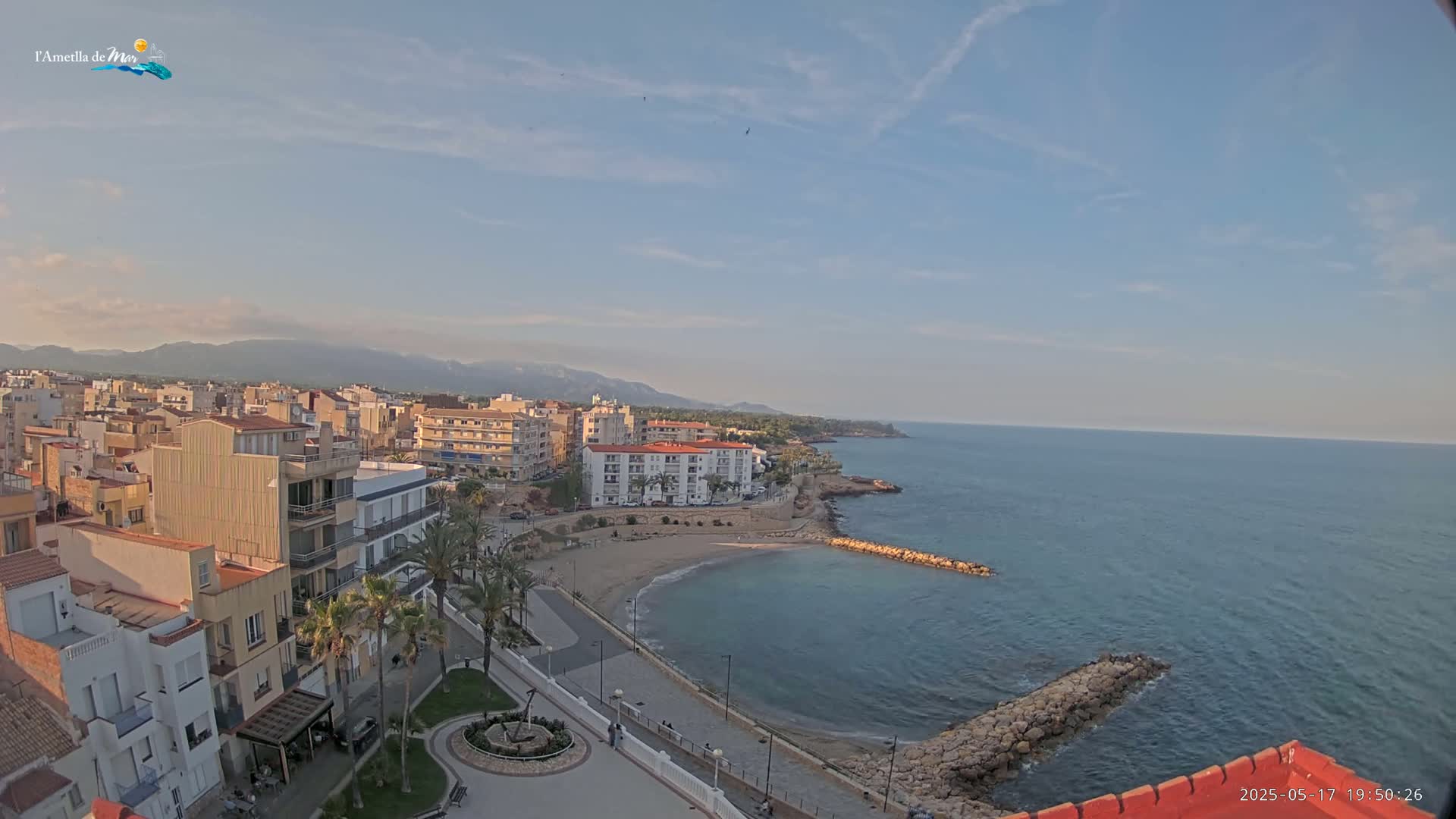 An aerial view of a coastal town on a partly sunny day, showing buildings, a small beach, and calm blue water extending to distant mountains.