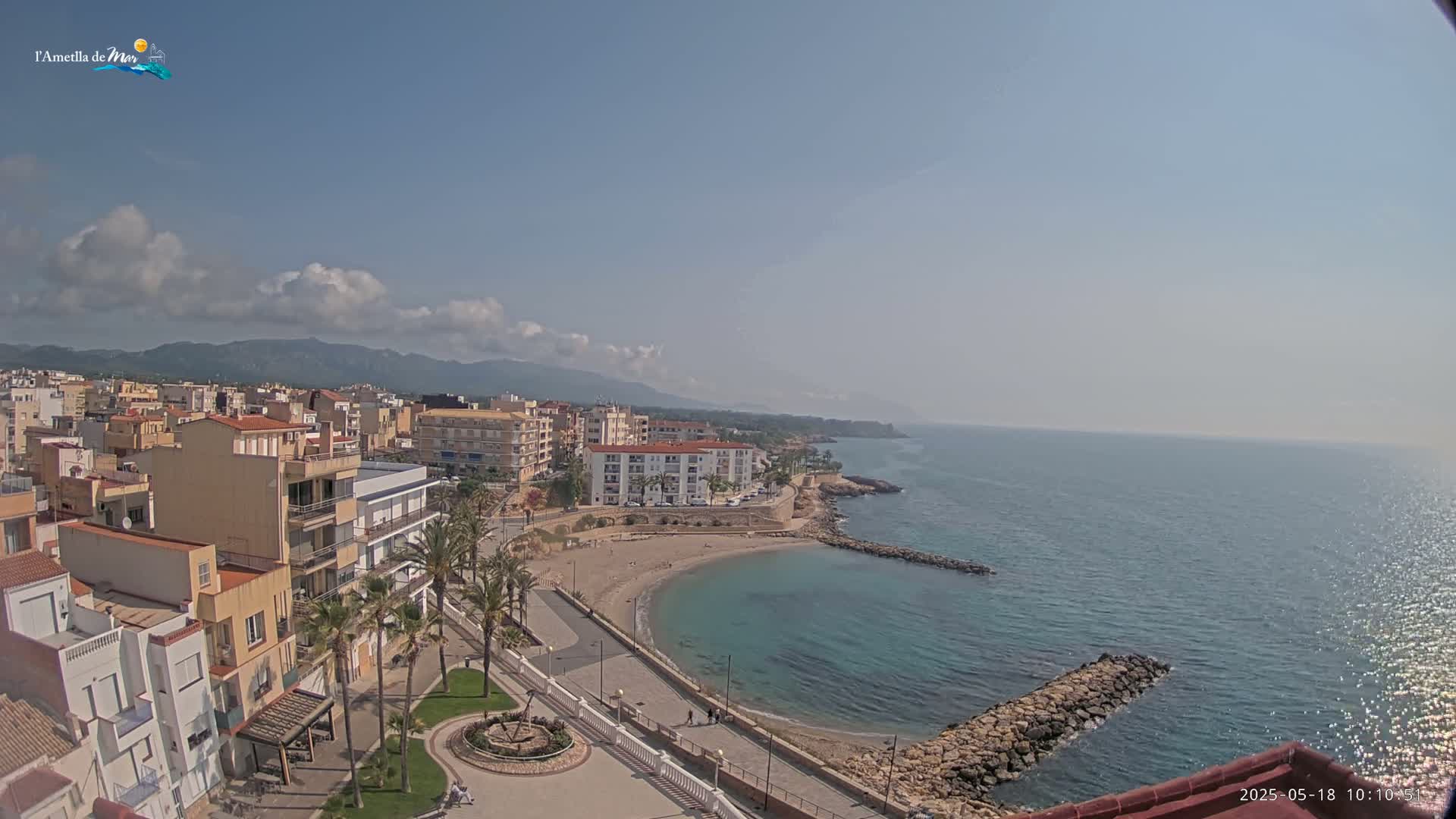 A coastal town with buildings and palm trees overlooks a calm, blue ocean cove under a partly cloudy sky.