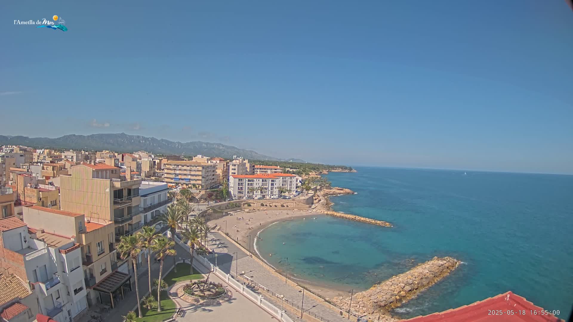 A coastal town with buildings, palm trees, a sandy beach cove, and calm blue ocean under a clear blue sky.