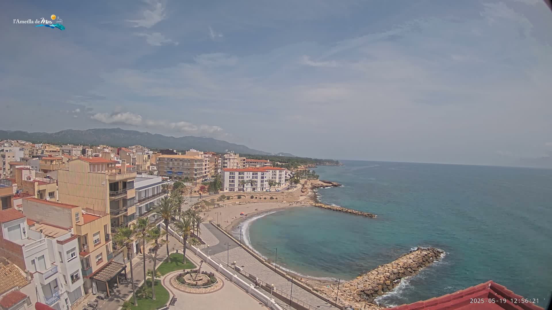 A mostly sunny day reveals a coastal town with buildings, palm trees, a small beach, and a calm turquoise sea backed by mountains in the distance.