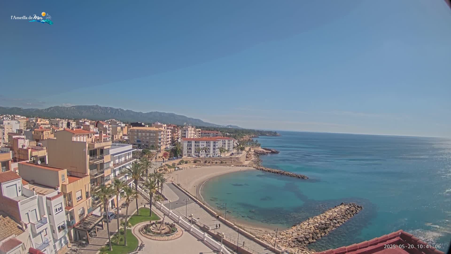 A coastal town with buildings, palm trees, and a small, crescent-shaped beach is visible on a sunny day with clear blue skies and calm turquoise waters.