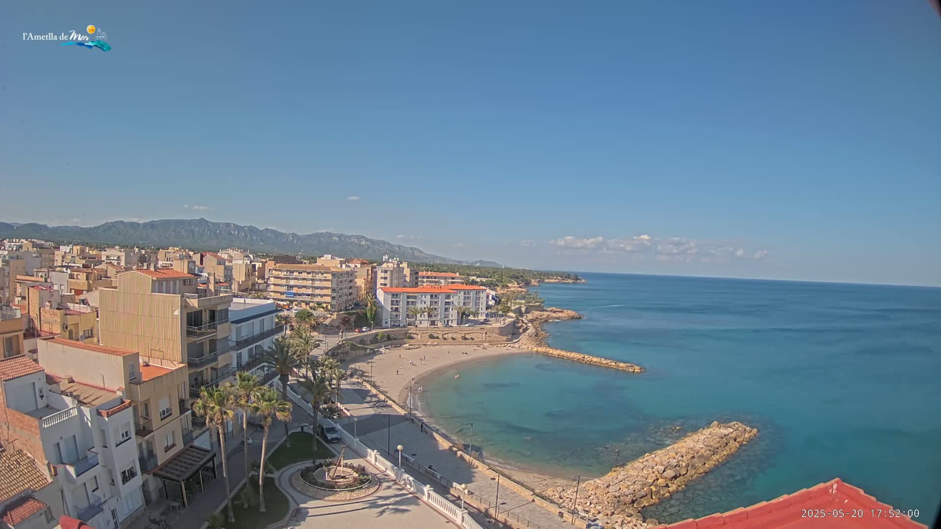 A coastal town with buildings and a sandy beach curving into a turquoise bay under a clear blue sky with a few clouds.
