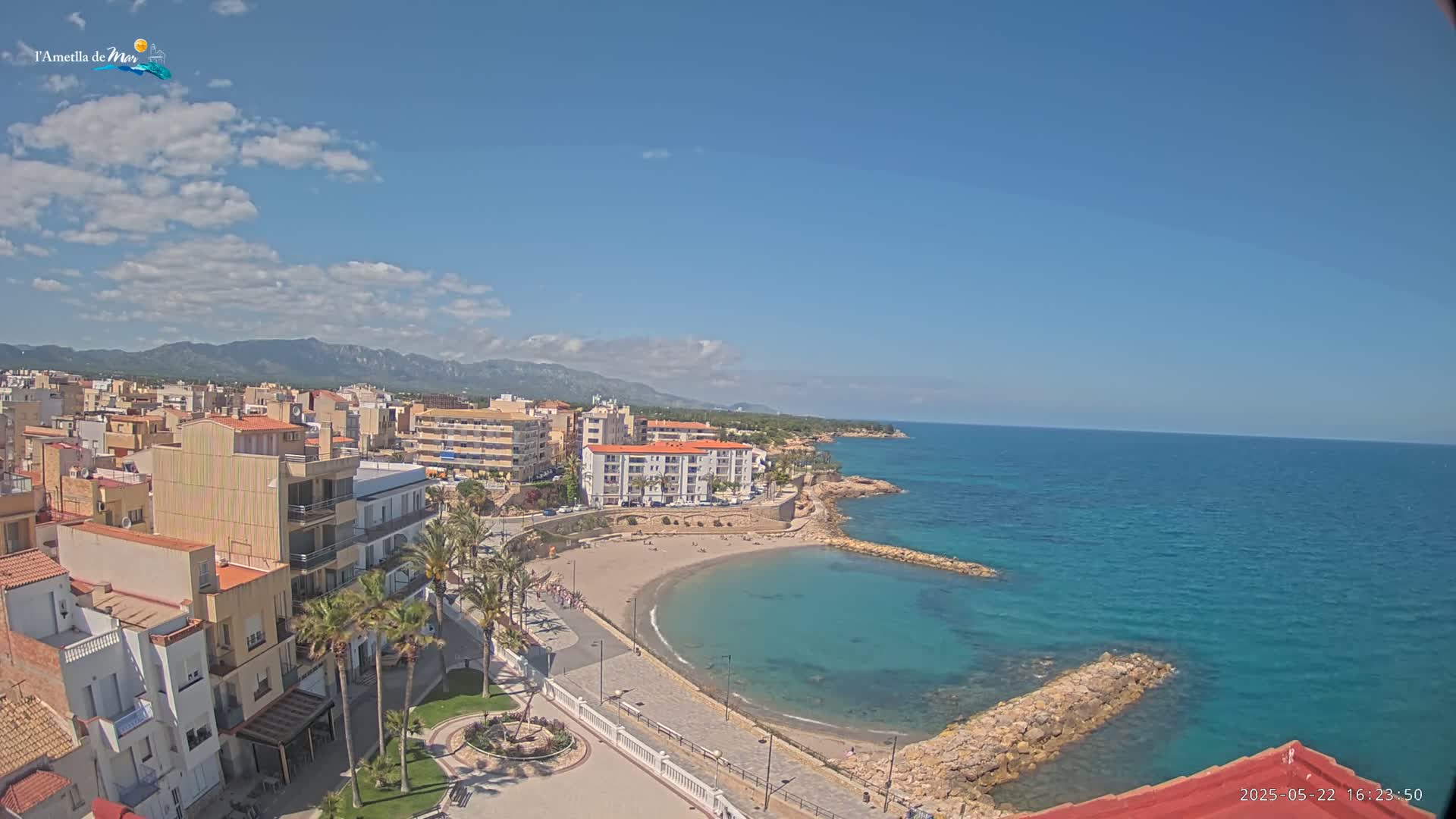 An aerial view shows a coastal town under a partly cloudy sky, featuring buildings, a small crescent-shaped beach, and a calm, turquoise sea.