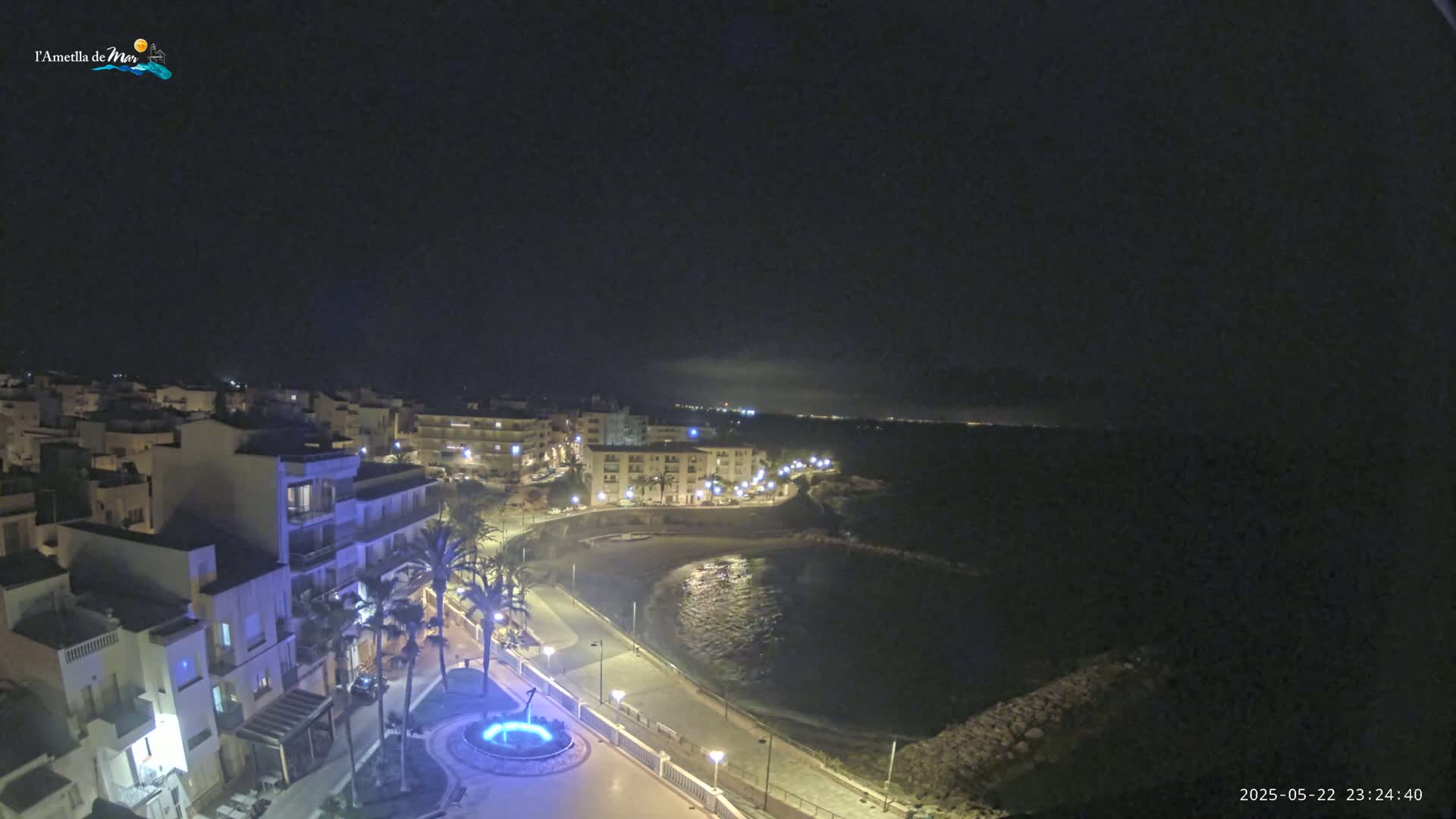 A nighttime coastal town scene shows a crescent-shaped bay with calm water, a paved waterfront path lined with palm trees and lights, and buildings illuminated against a dark sky.