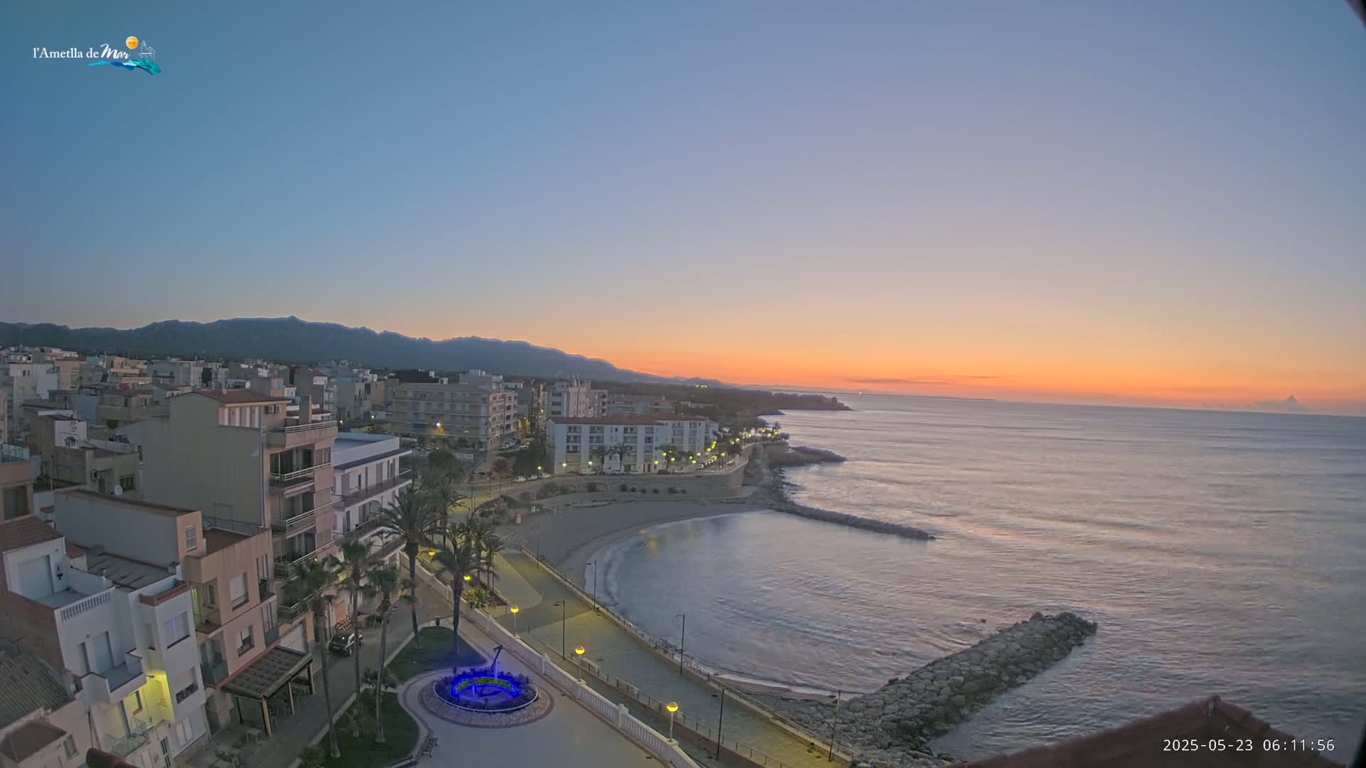 A coastal town at sunrise, with calm ocean waves gently lapping the shore, is seen from an elevated viewpoint.