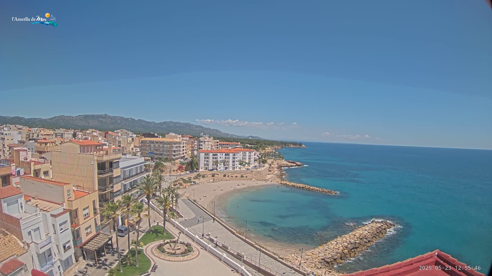 An aerial view shows a coastal town on a sunny day, with a small sandy cove and turquoise water extending to a calm ocean under a clear blue sky.