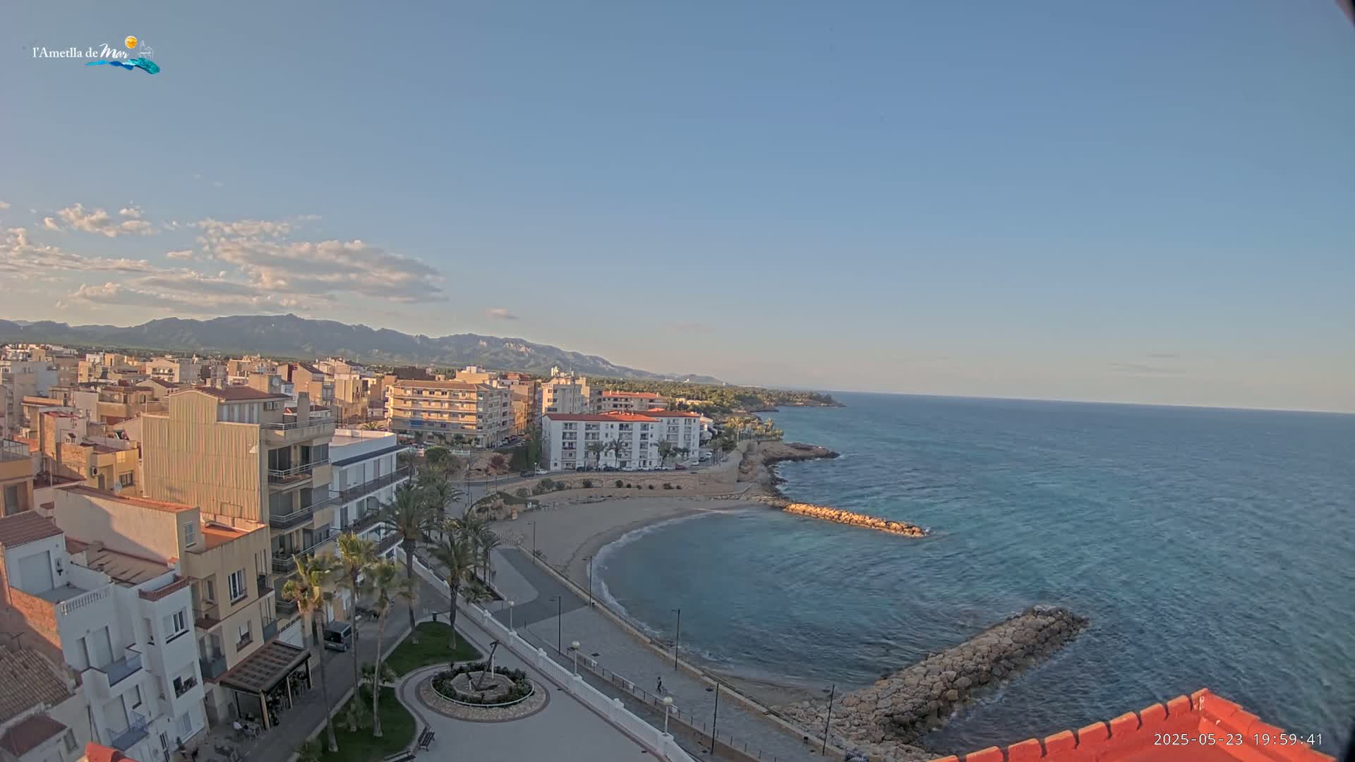 A coastal town with buildings and a small beach is visible under a partly cloudy sky, overlooking a calm sea and distant mountains.