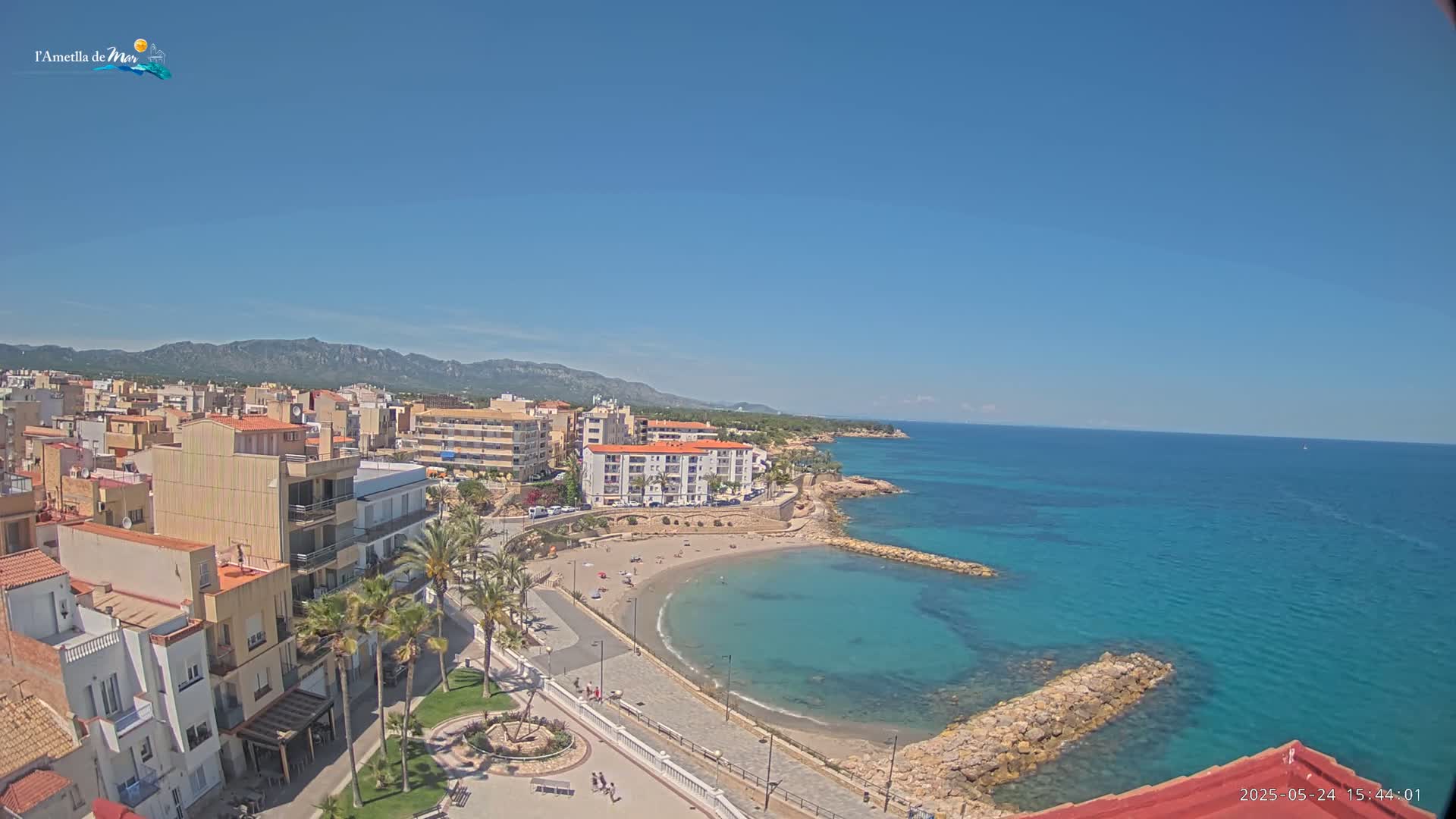 An elevated view shows a coastal town on a sunny day, with a small, crescent-shaped beach and clear, turquoise water.