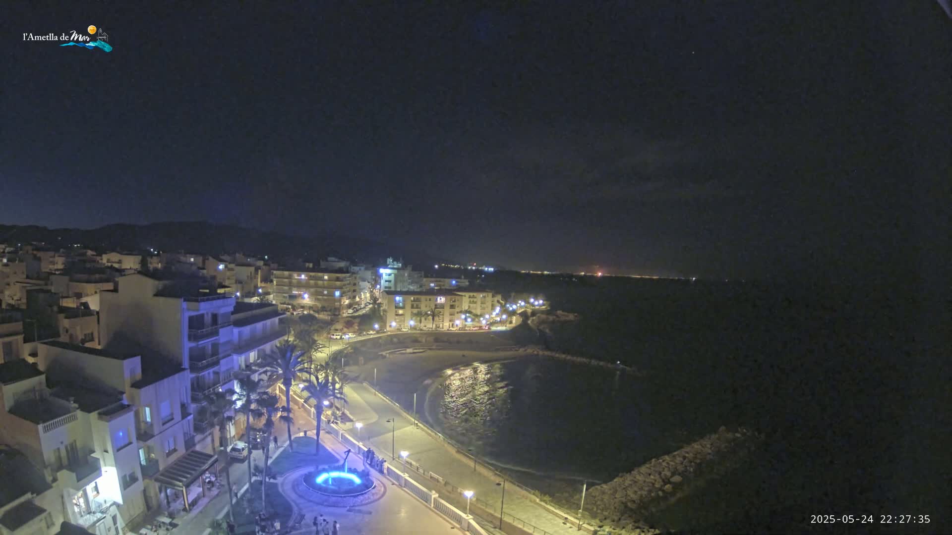 A nighttime, coastal town scene shows a crescent-shaped bay with a waterfront promenade lined with buildings and palm trees under a clear, dark sky.