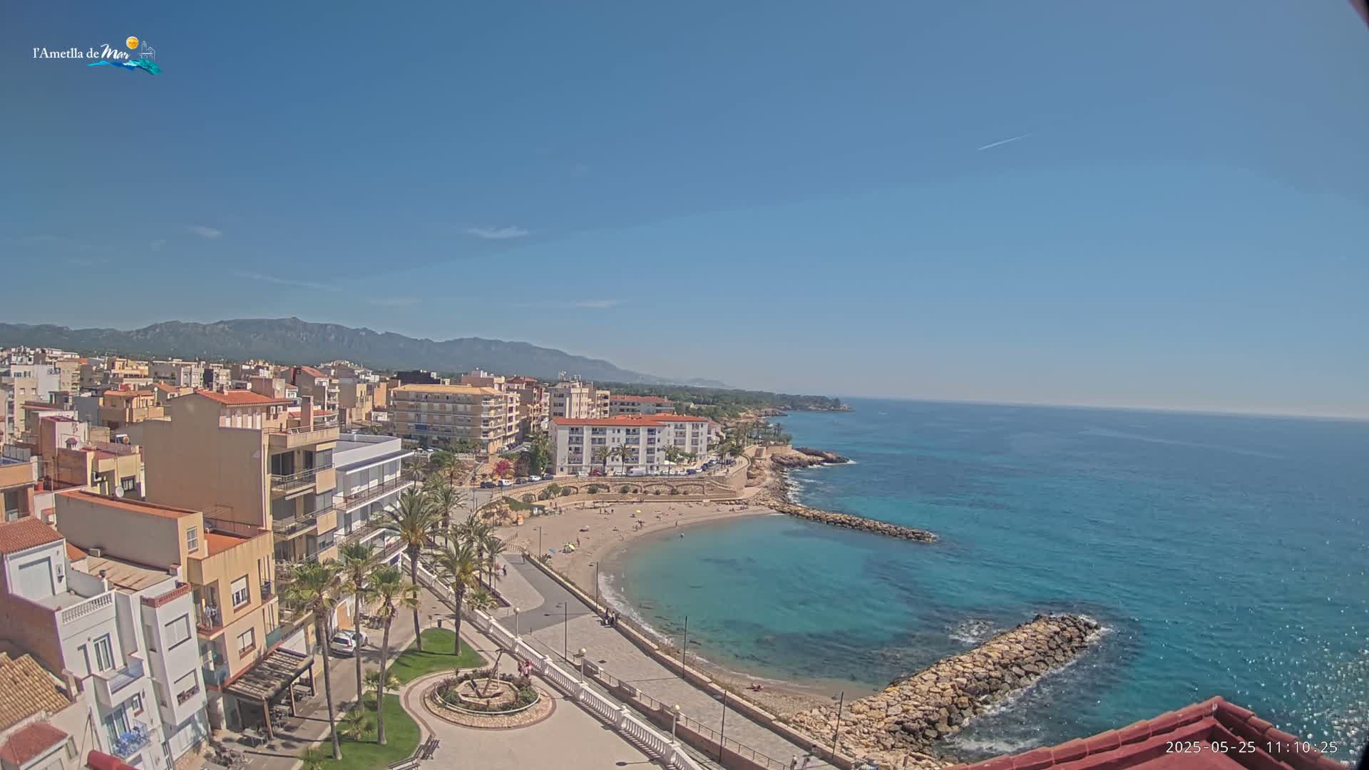 An aerial view shows a coastal town on a sunny day, with a small, sandy cove and calm, turquoise water stretching to the horizon, backed by buildings and mountains.
