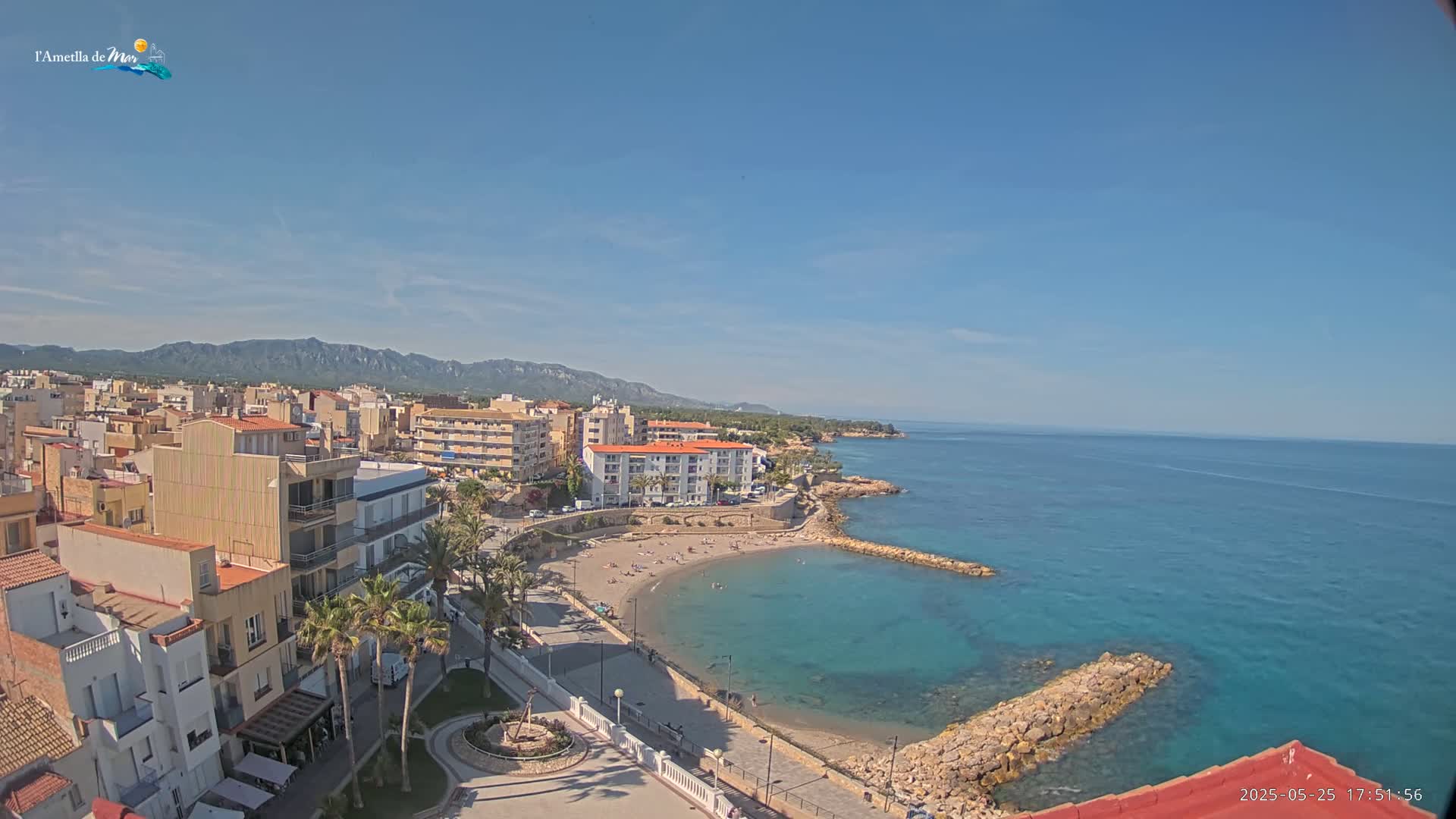 An elevated view shows a coastal town on a sunny day, with a small sandy beach curving into a calm, teal ocean, backed by buildings and mountains.