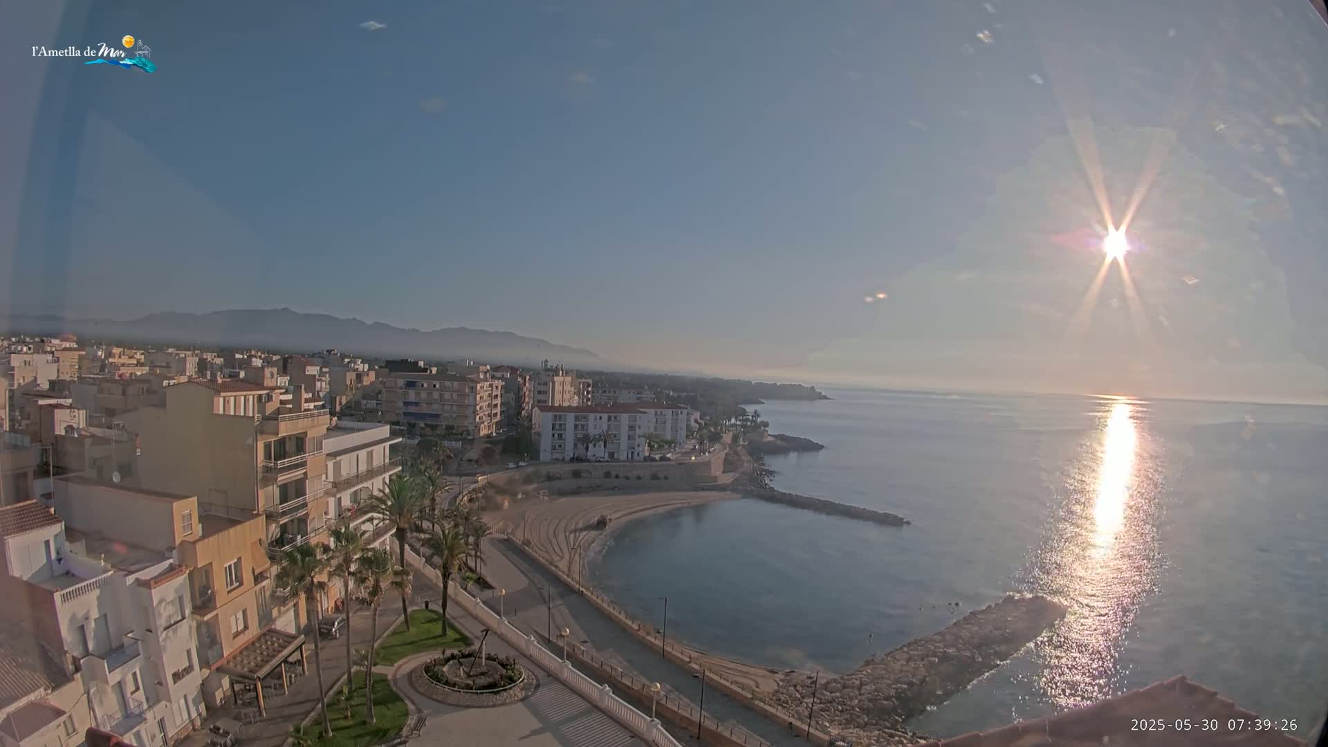 A coastal town with buildings, palm trees, and a small sandy cove is seen on a sunny morning with a clear sky and calm sea.