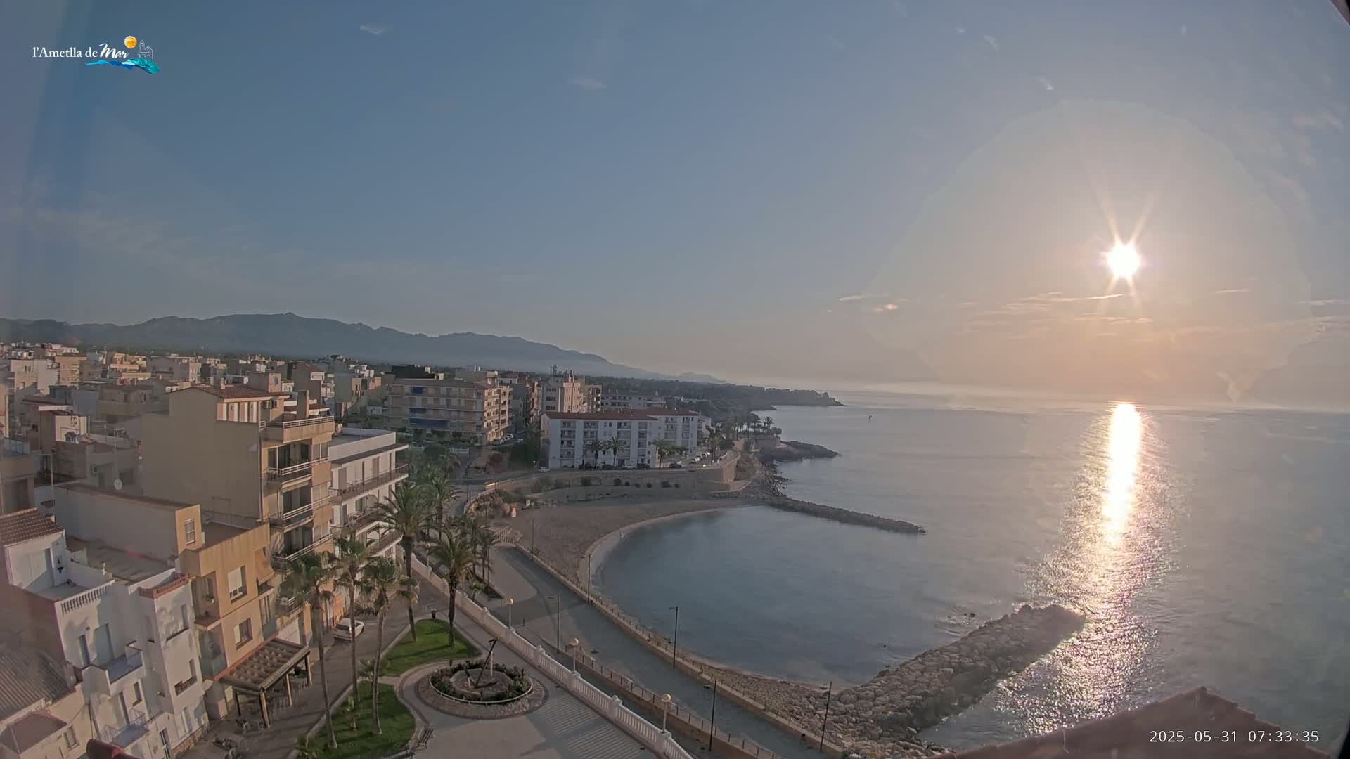 A coastal town at sunrise, with calm waters reflecting the bright sun, is seen from an elevated vantage point.
