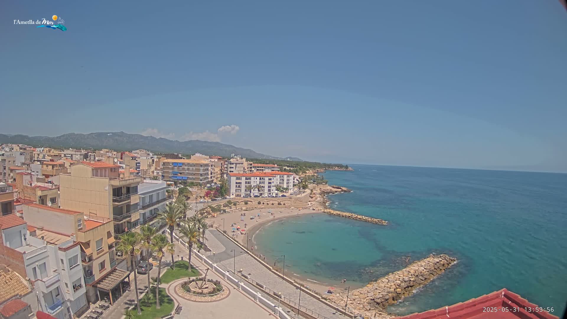 An aerial view shows a coastal town on a sunny day with a small, curved beach and calm, clear turquoise water.