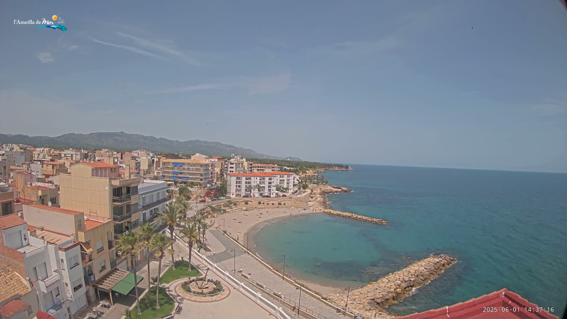 An elevated view shows a coastal town with buildings, palm trees, a small sandy beach, and calm, clear turquoise water under a mostly clear sky.