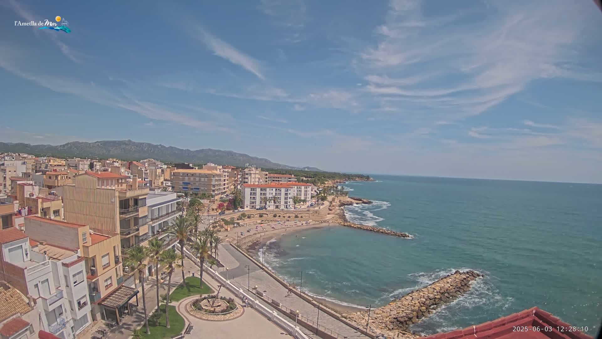 A coastal town with buildings, a small beach with people, and a calm ocean under a mostly sunny sky.