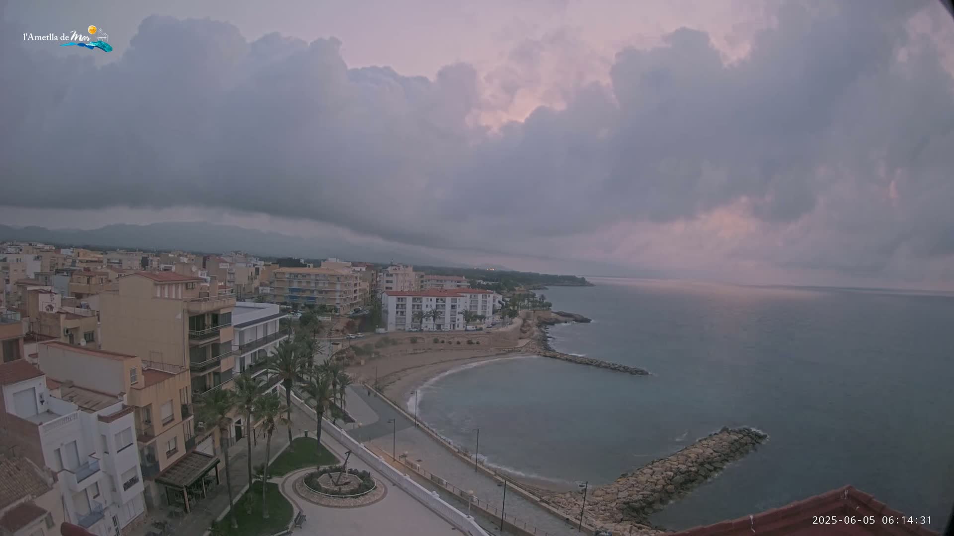 A coastal town with low buildings and palm trees is partially obscured by a large, dark cloud formation under an overcast sky, overlooking a calm sea.