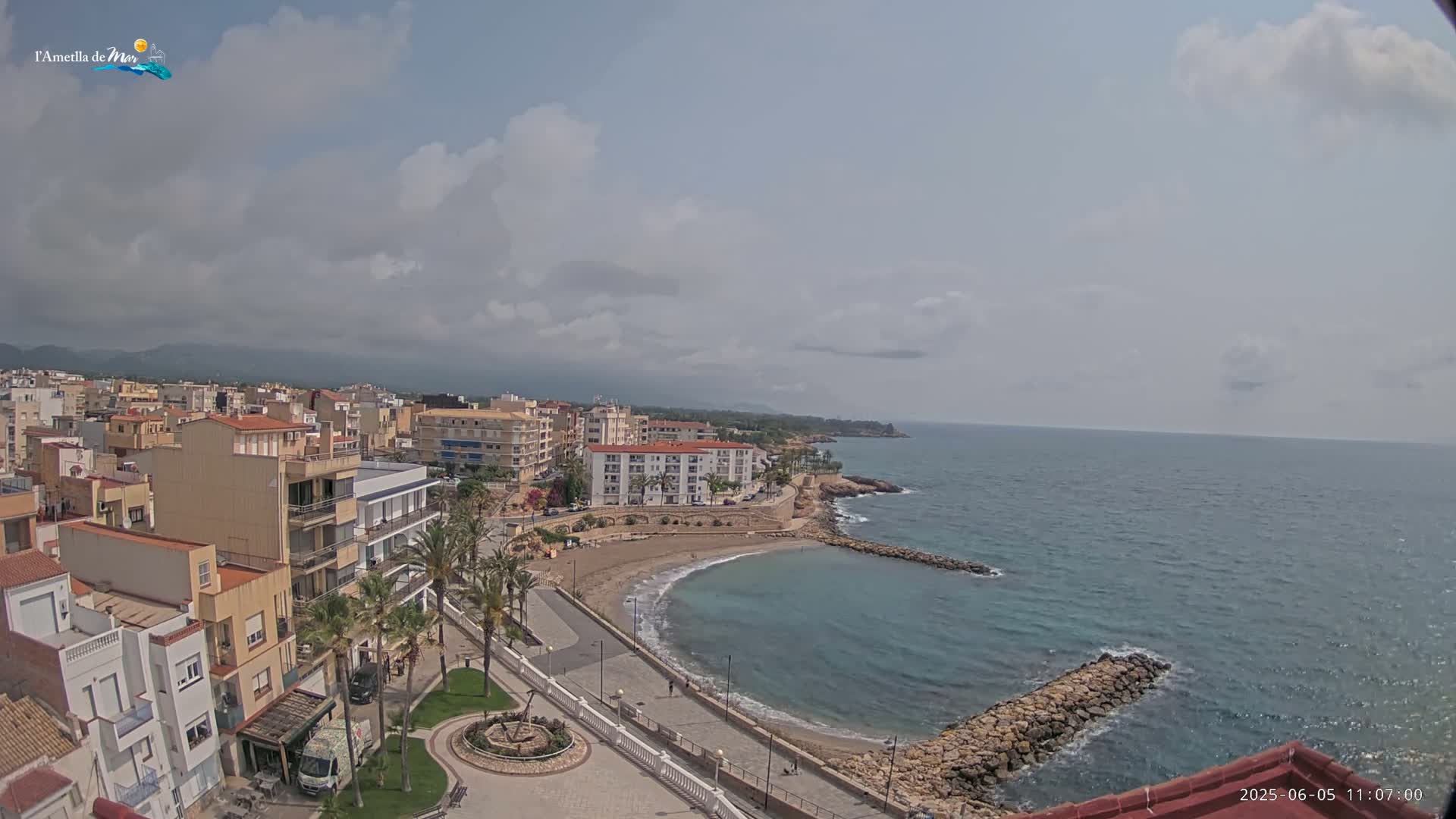 A partly cloudy day reveals a coastal town with buildings clustered near a calm, blue sea featuring a small, sandy cove.