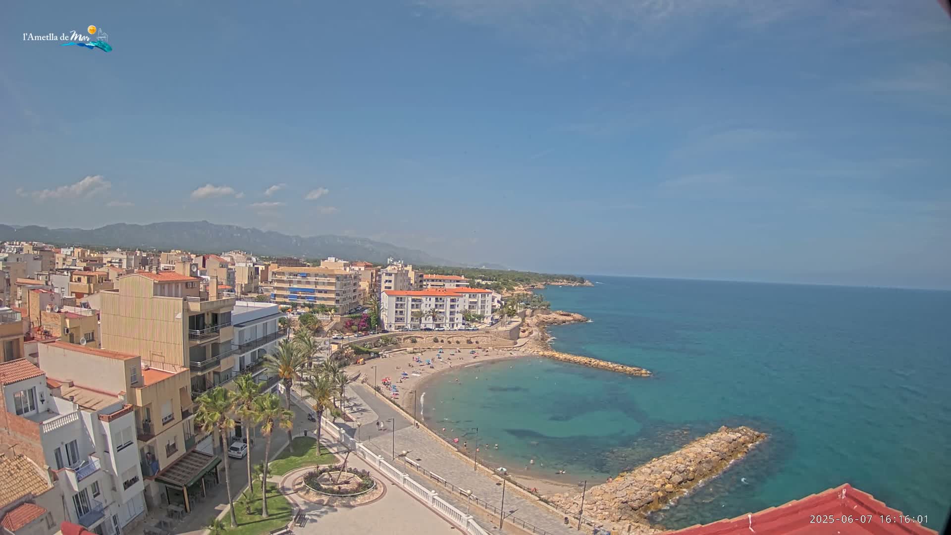 A sunny coastal town with a small sandy beach and calm, clear turquoise water, backed by buildings and mountains under a mostly clear sky.