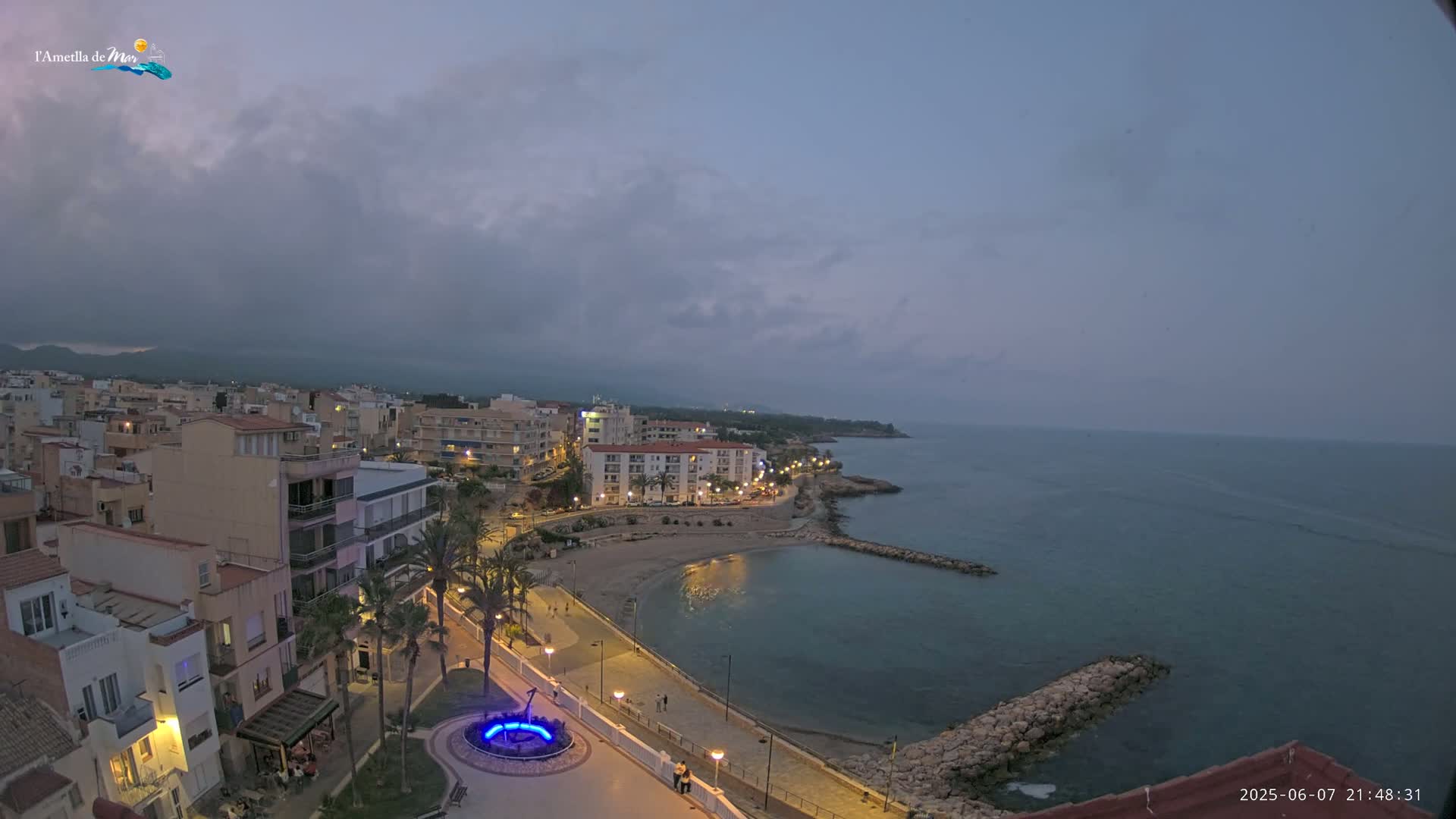 A coastal town at dusk under a cloudy sky, with a calm sea and a small beach, is illuminated by streetlights.