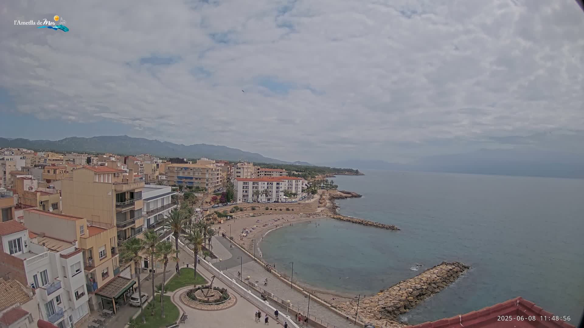 An aerial view shows a coastal town with buildings, a small beach with people, and a calm sea under a mostly cloudy sky.