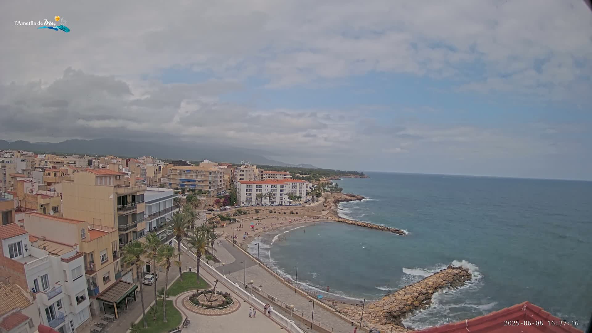 A partly cloudy day reveals a coastal town with a small sandy beach and calm ocean waters partially enclosed by rock breakwaters.