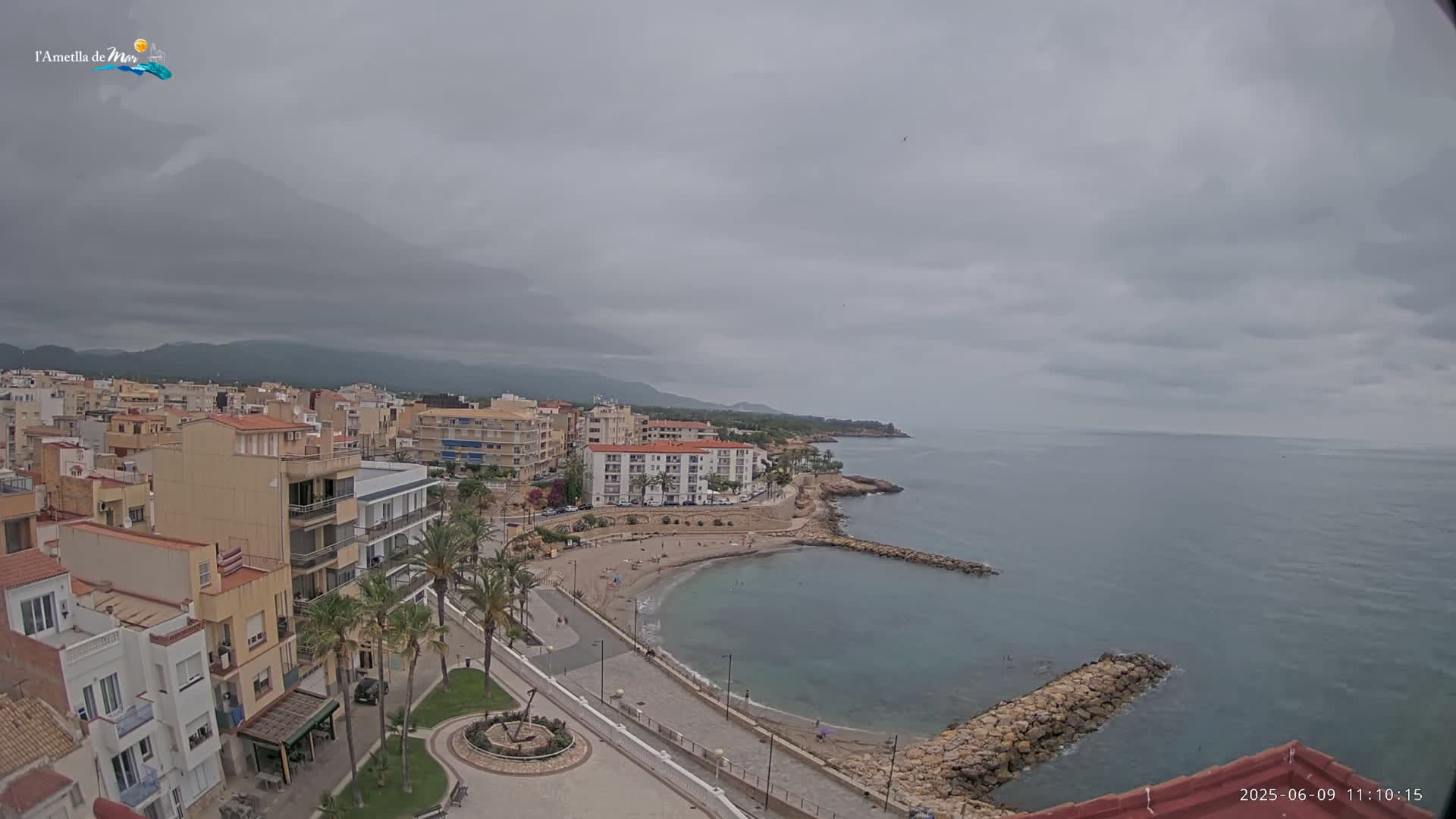 An overcast day reveals a coastal town with multi-story buildings, a small sandy beach curving into a calm bay, and a rocky breakwater extending into the sea.