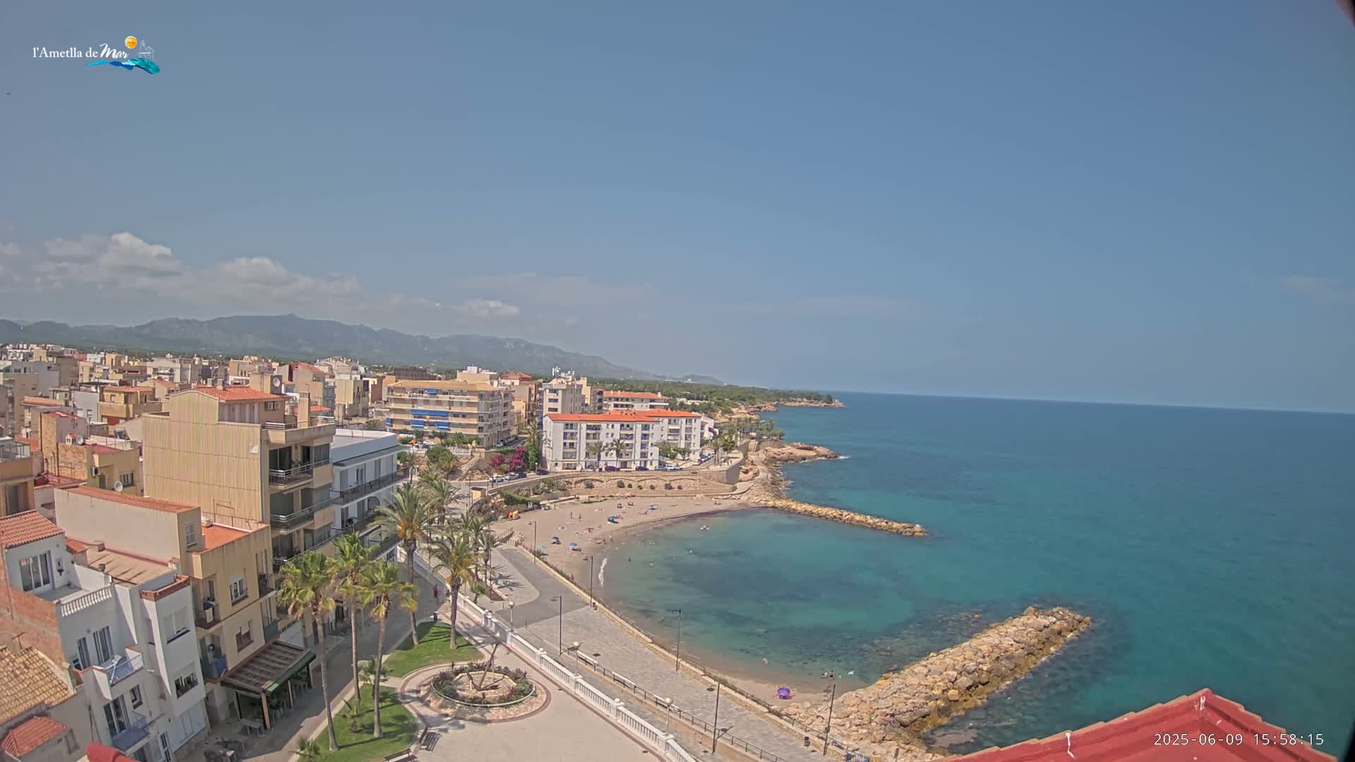 An aerial view shows a coastal town on a sunny day with a small, sandy beach and calm, turquoise water, backed by buildings and distant mountains.