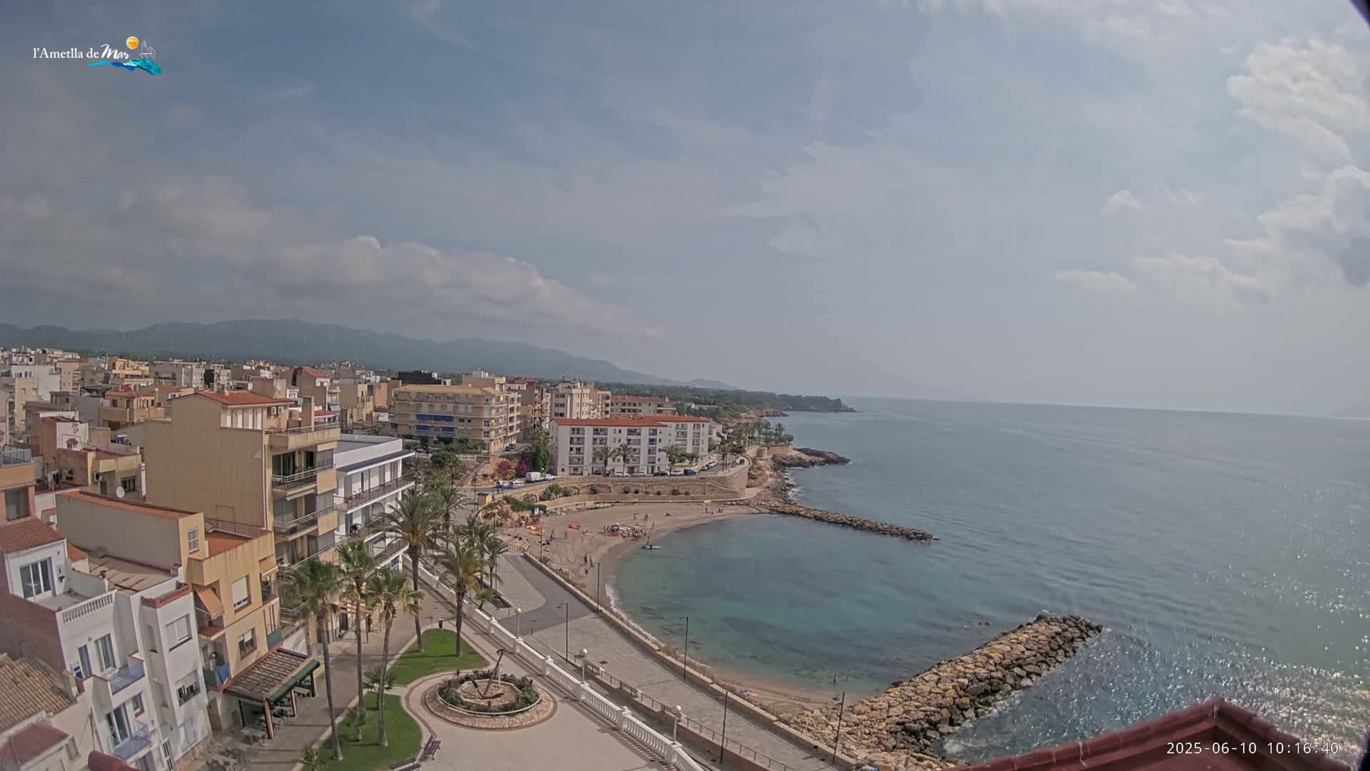 A partly cloudy day reveals a coastal town with a small sandy beach and calm turquoise water, backed by buildings and distant mountains.