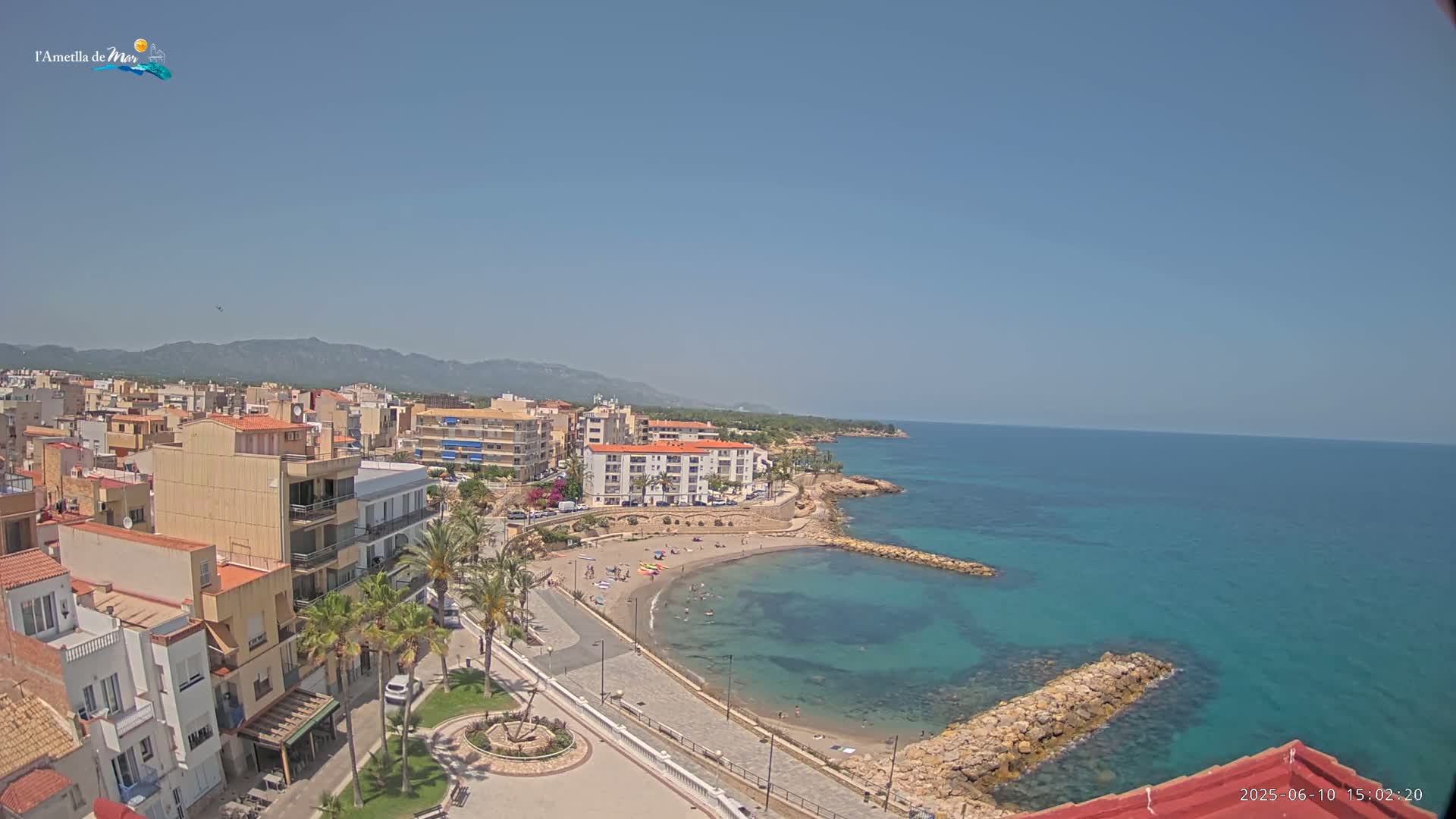 A coastal town with buildings, palm trees, and a small sandy beach nestled in a calm, turquoise bay under a clear blue sky.