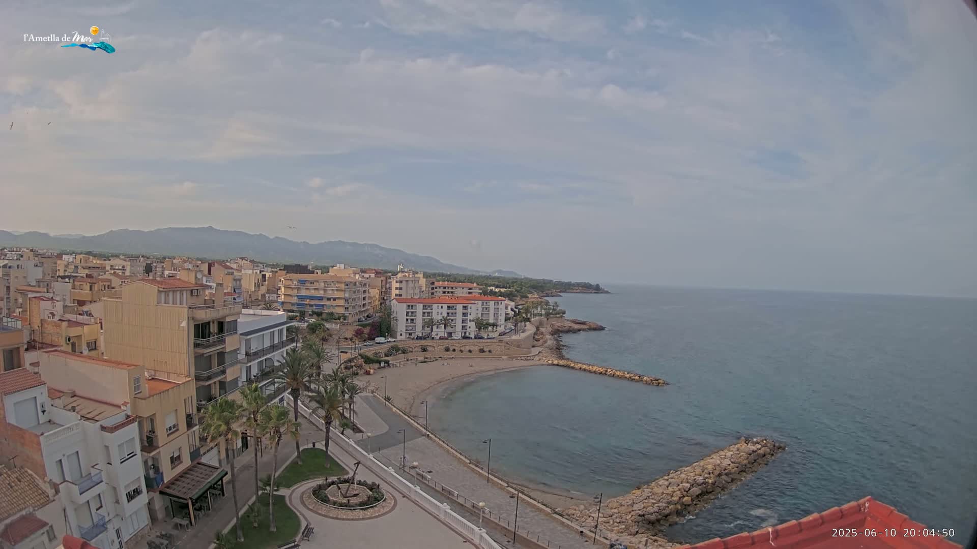 A partly cloudy day reveals a coastal town with buildings, palm trees, a small sandy beach, and a calm sea with rocky breakwaters.
