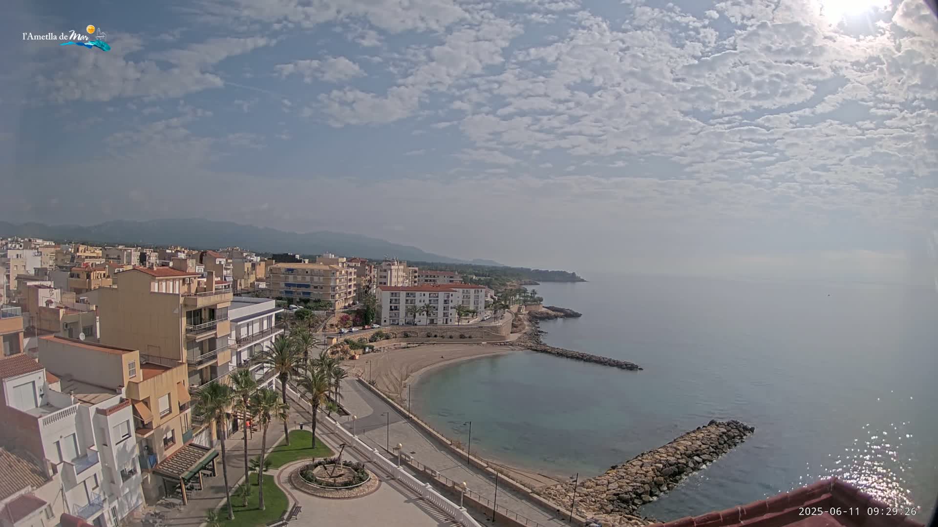 A coastal town with buildings, palm trees, a sandy beach curving into a calm bay under a partly cloudy sky.