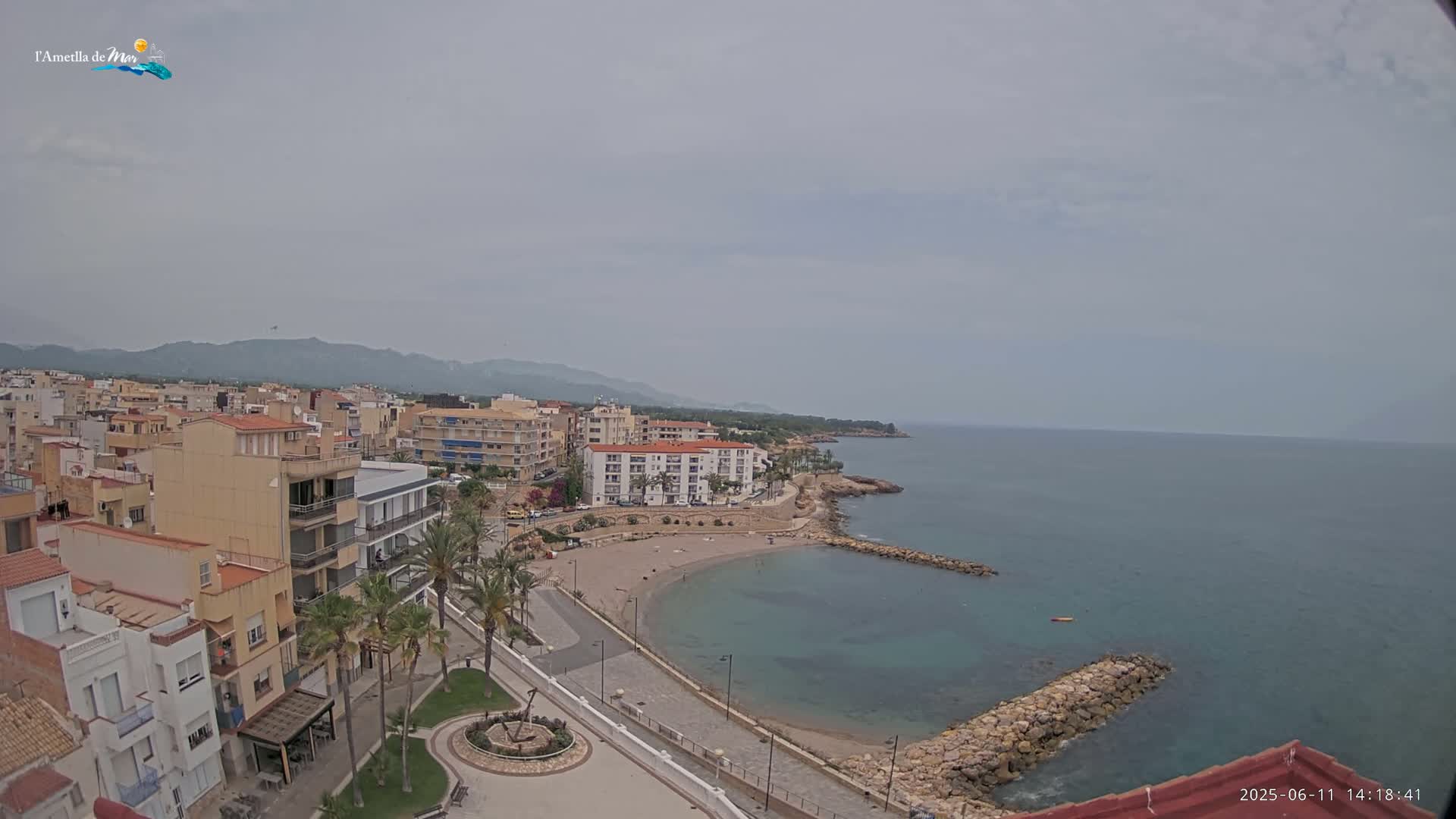 A coastal town with buildings and palm trees sits on a small, sandy cove under an overcast sky.