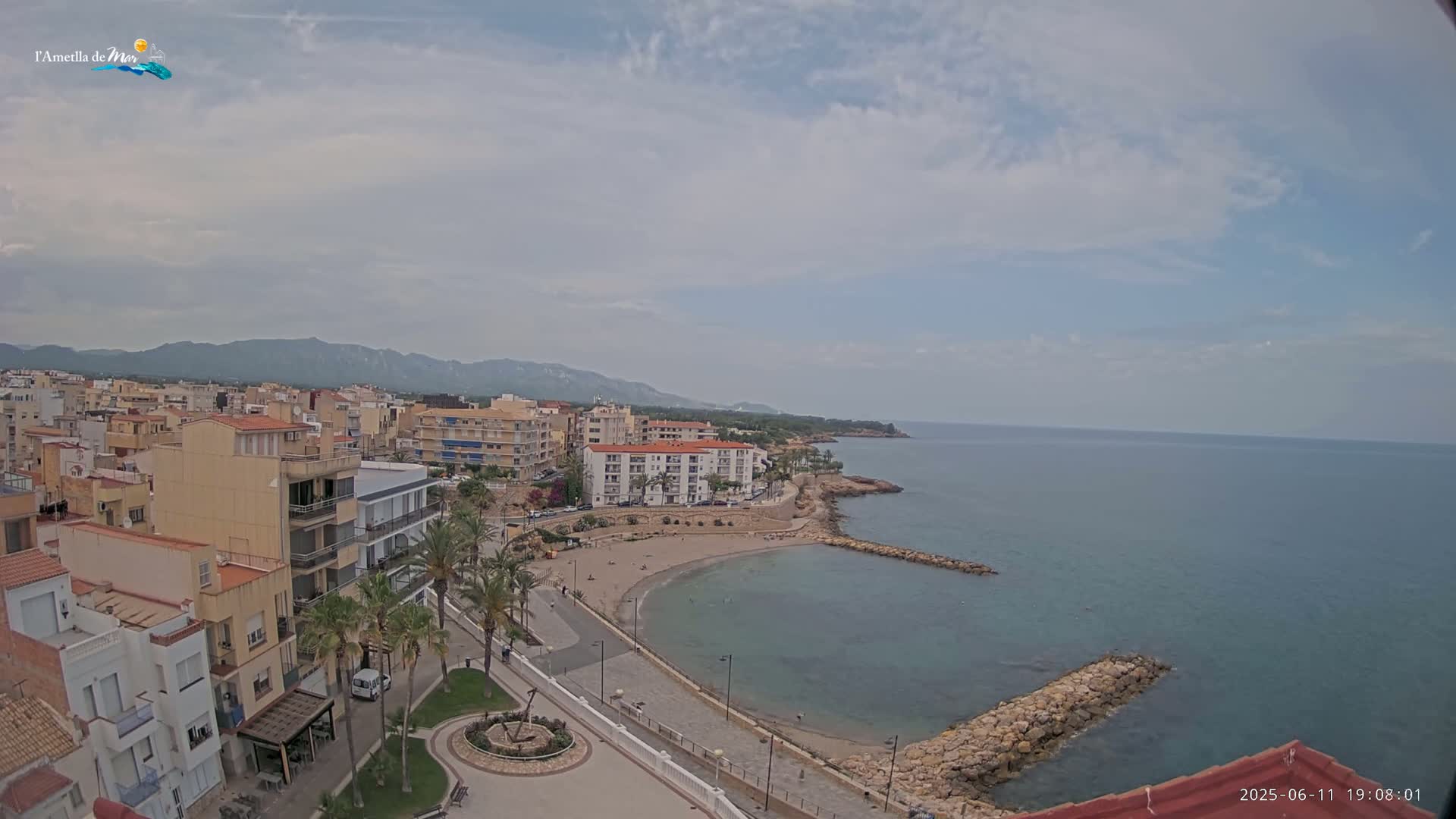 A coastal town with buildings, palm trees, and a small beach is visible under a partly cloudy sky.