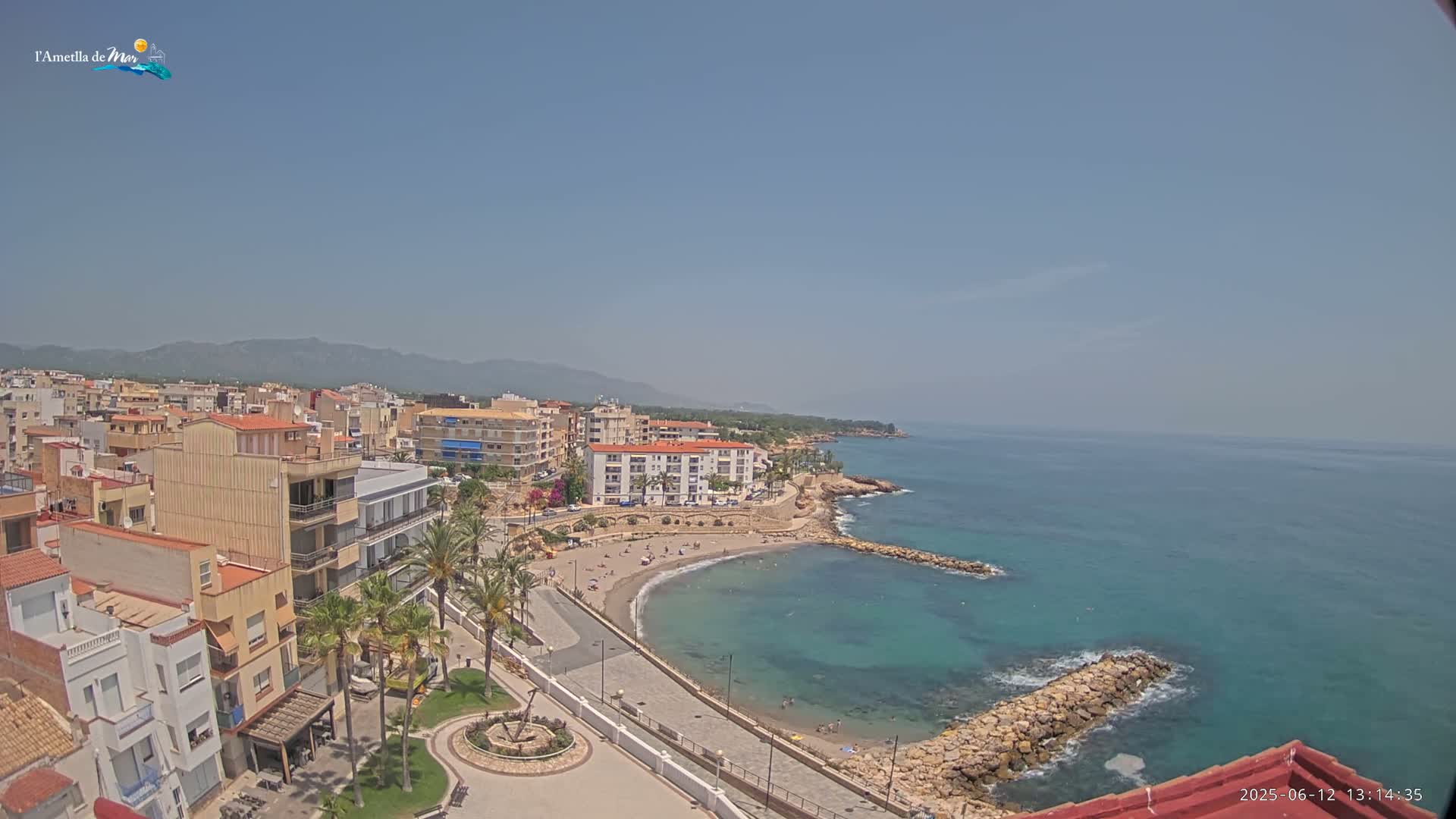 A coastal town with buildings, palm trees, and a small sandy beach with calm, clear turquoise water under a clear, sunny sky.